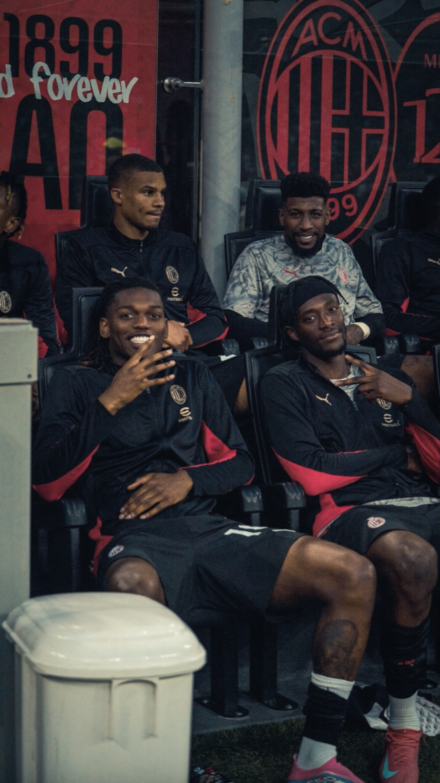 Group of soccer players sitting on bench, wearing black and red team jackets, in team dugout with AC Milan logo and banners in background.