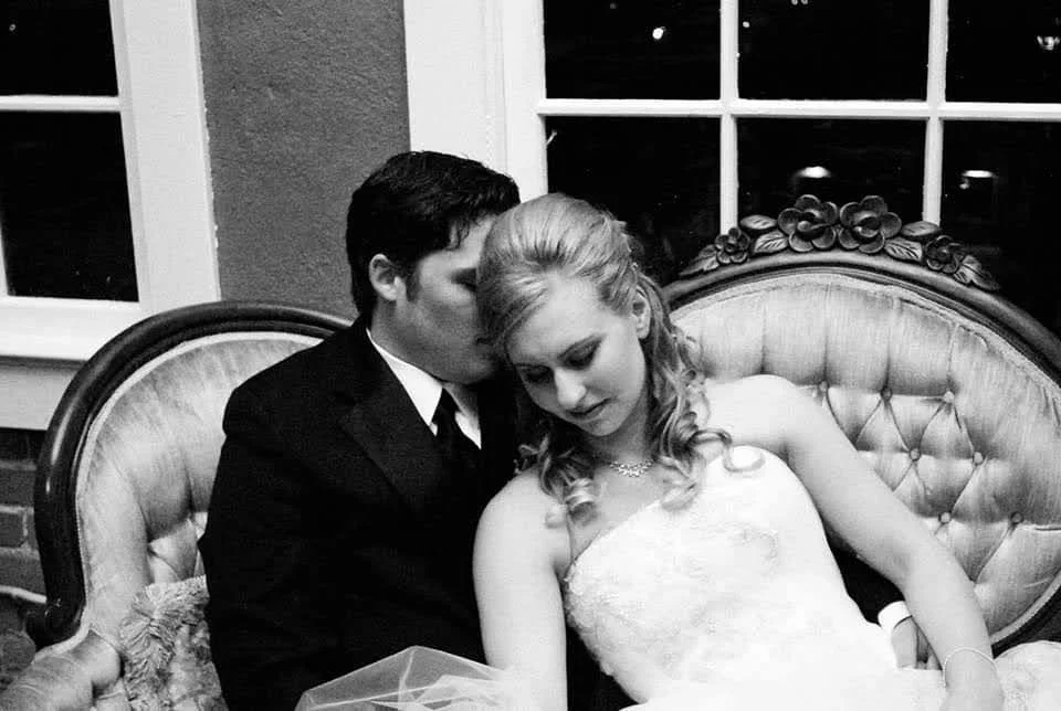 A man in a tuxedo kisses a woman in a wedding dress on the head while sitting on a vintage-style sofa.