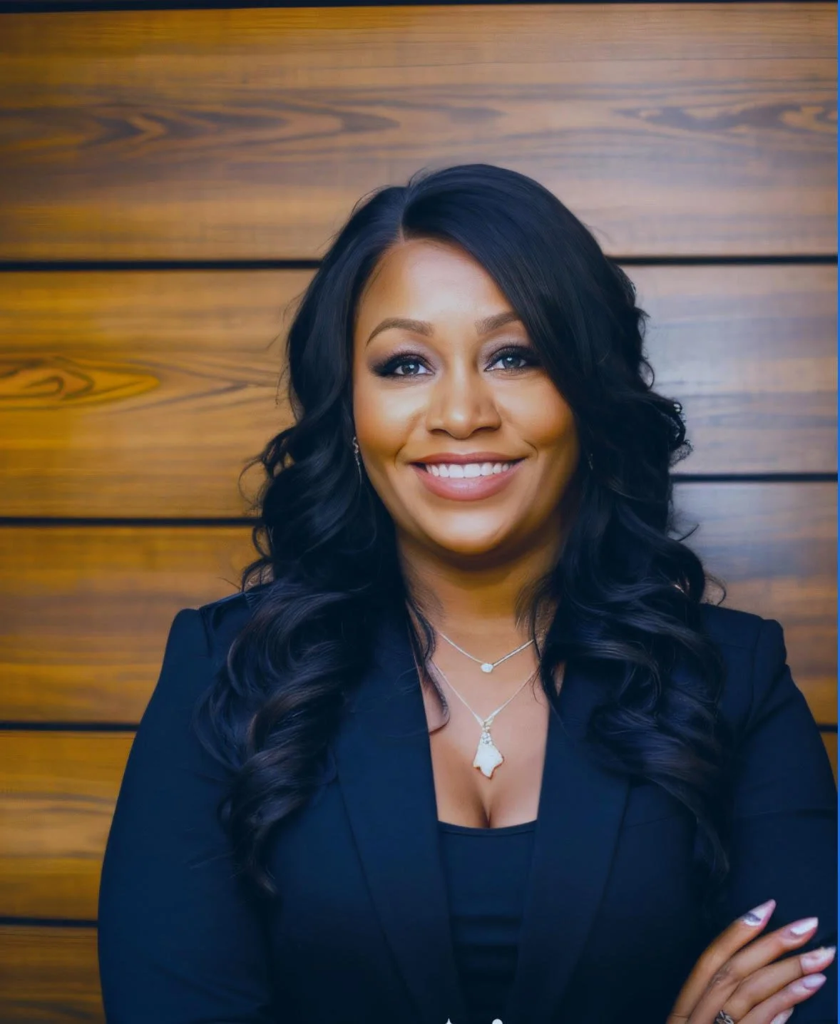 A woman with long, wavy black hair, wearing a black blazer and jewelry, smiling with her arms crossed in front of a wooden-paneled wall.