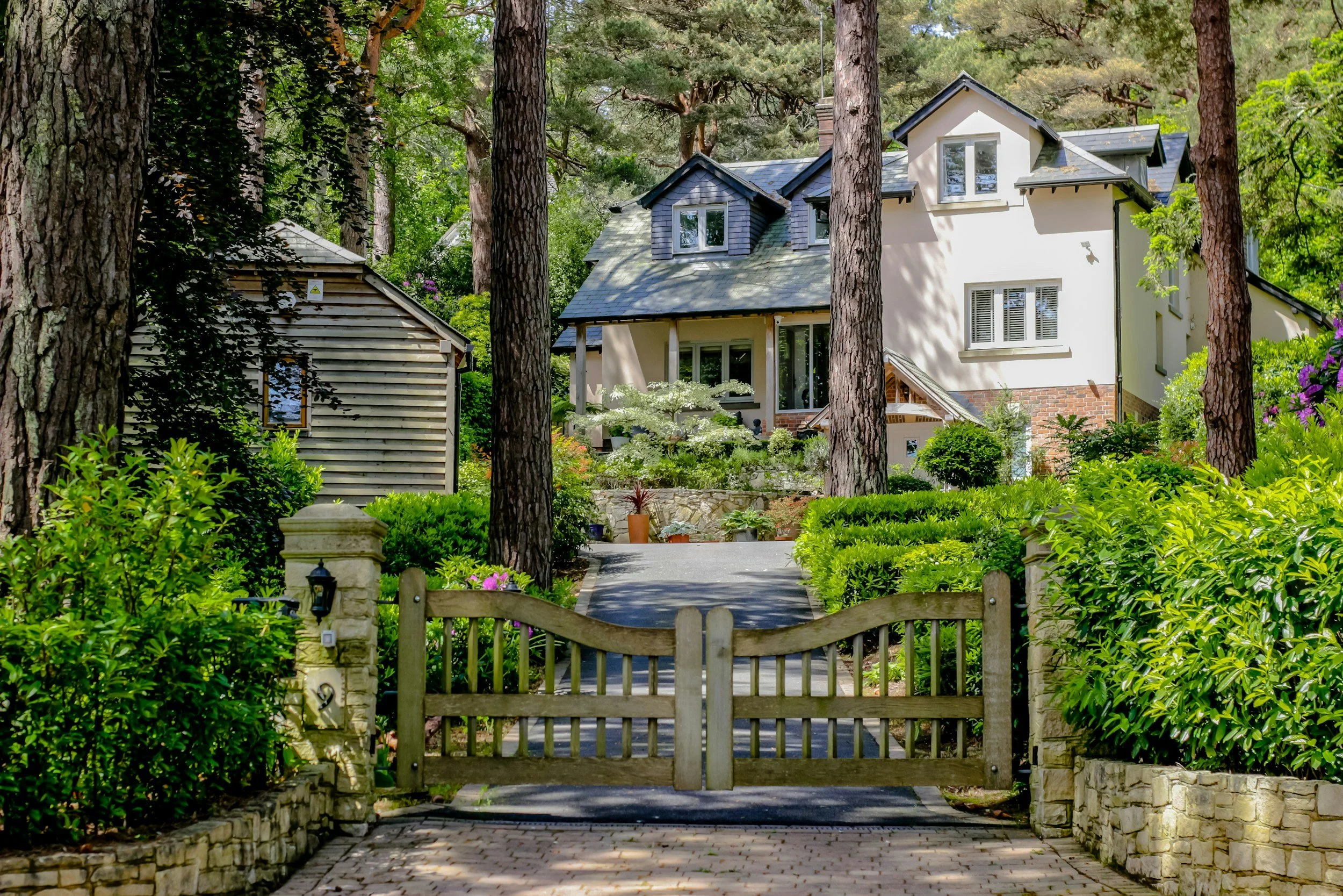 A view of a house in a lush, green forested area with tall trees, a pathway leading to the house, and a wooden gate at the front.