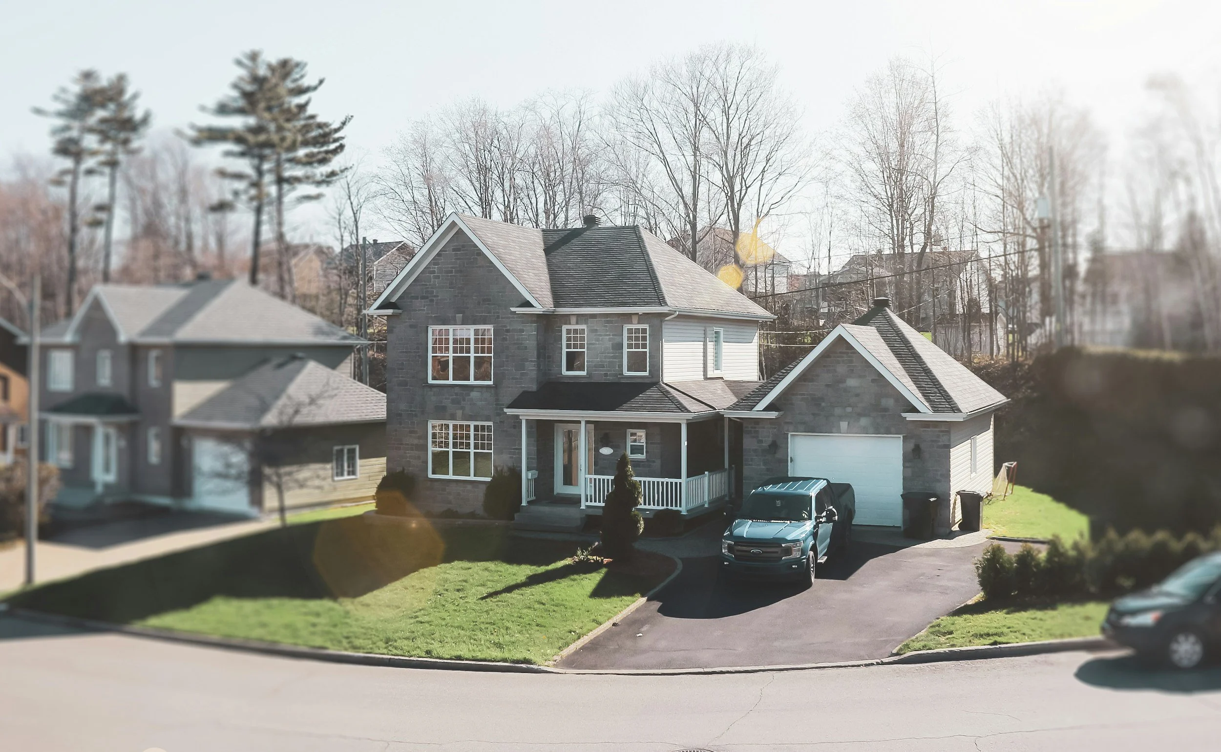A two-story house with gray stone exterior and a front porch, a black truck parked in the driveway, and neighboring houses visible in the background.