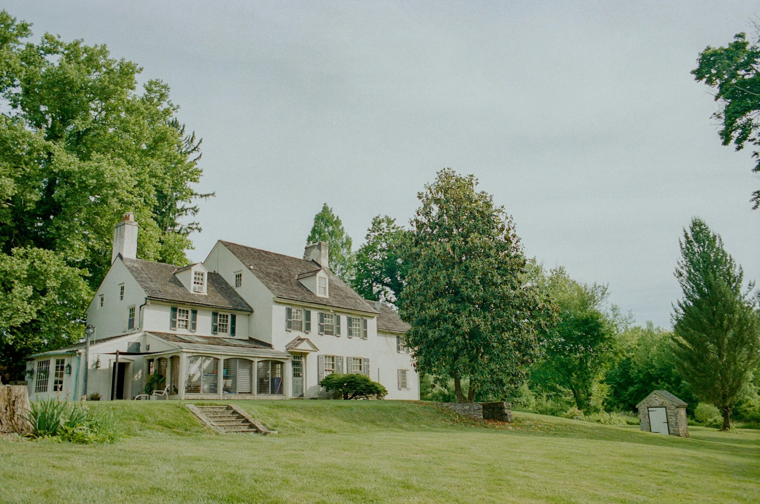 A white, two-story house with multiple chimneys, surrounded by green trees and a lawn.