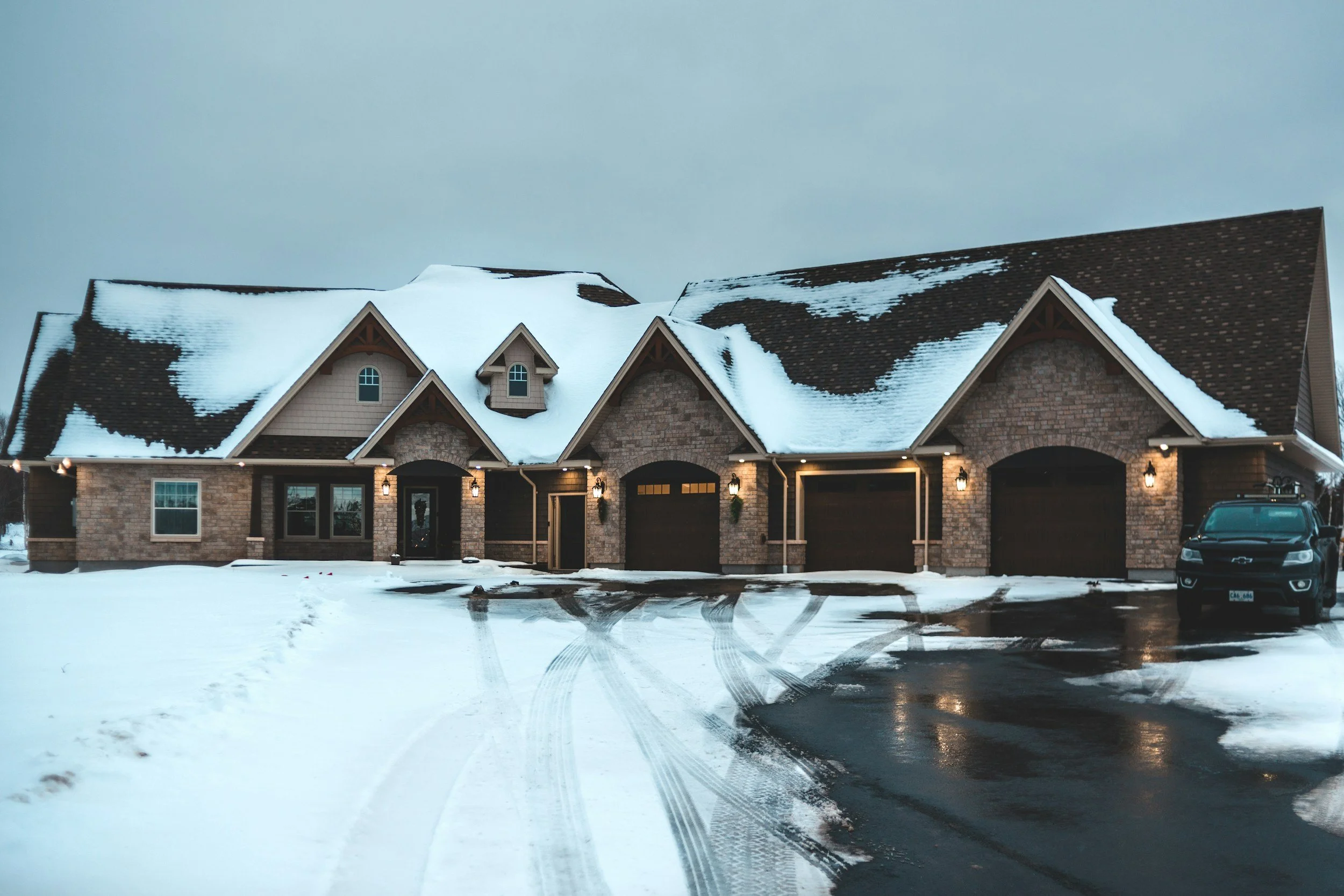 Large brick house with snow-covered roof and driveway, black garage doors, outdoor lighting, and a car parked outside during winter.