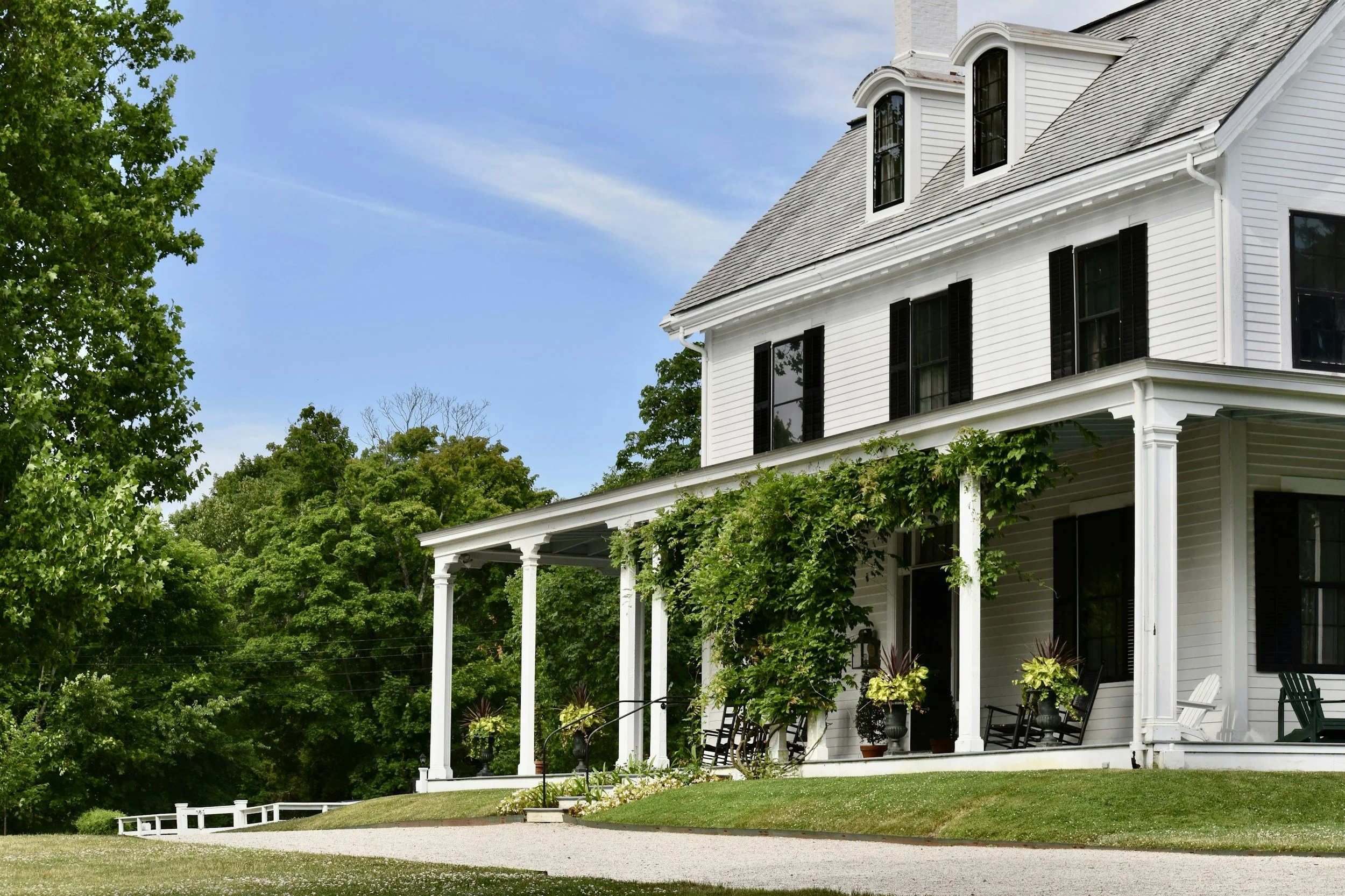 White house with black shutters, porch with chairs, surrounded by green trees and grass, blue sky.