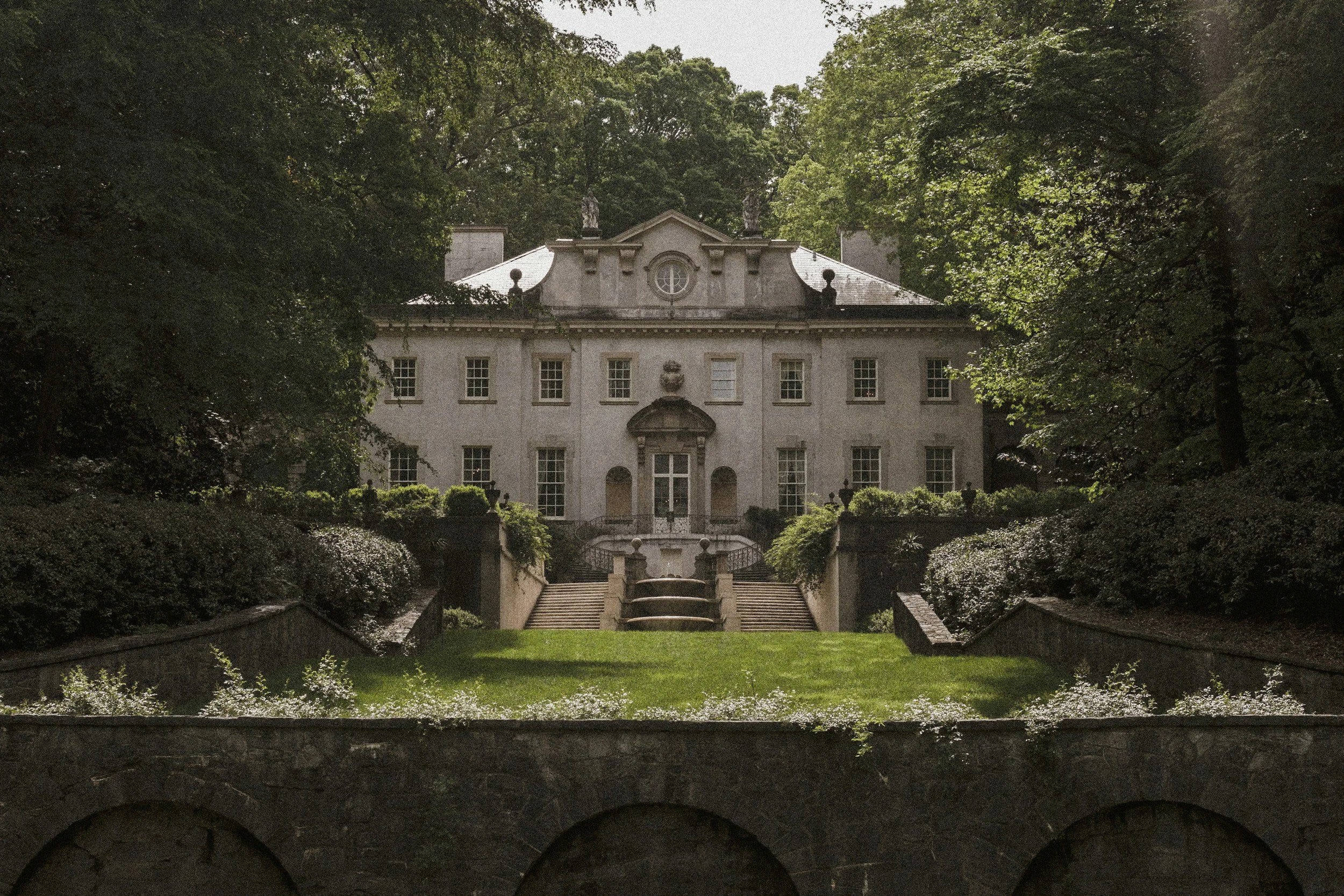 A large historic mansion with classical architecture, surrounded by lush greenery, with garden stairs leading up to the building