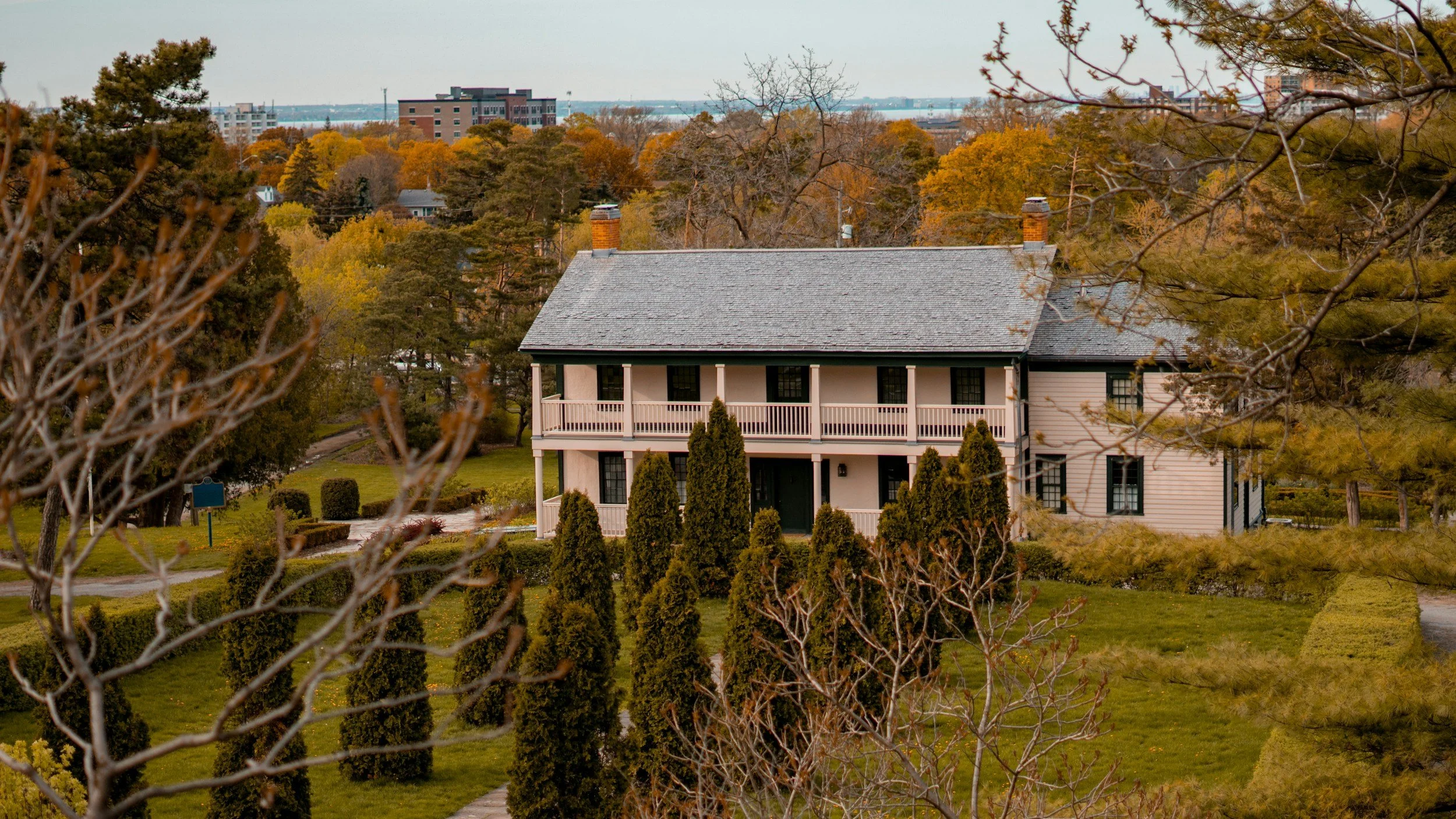 A two-story house with a gray roof, surrounded by trees and bushes, in an autumn landscape.