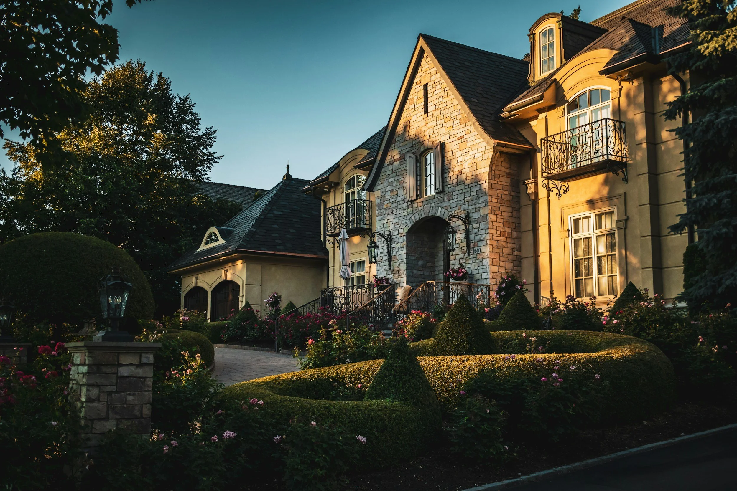 A large, elegant house with a stone and stucco exterior, surrounded by a well-maintained garden with trimmed bushes and pink flowers, during sunset.