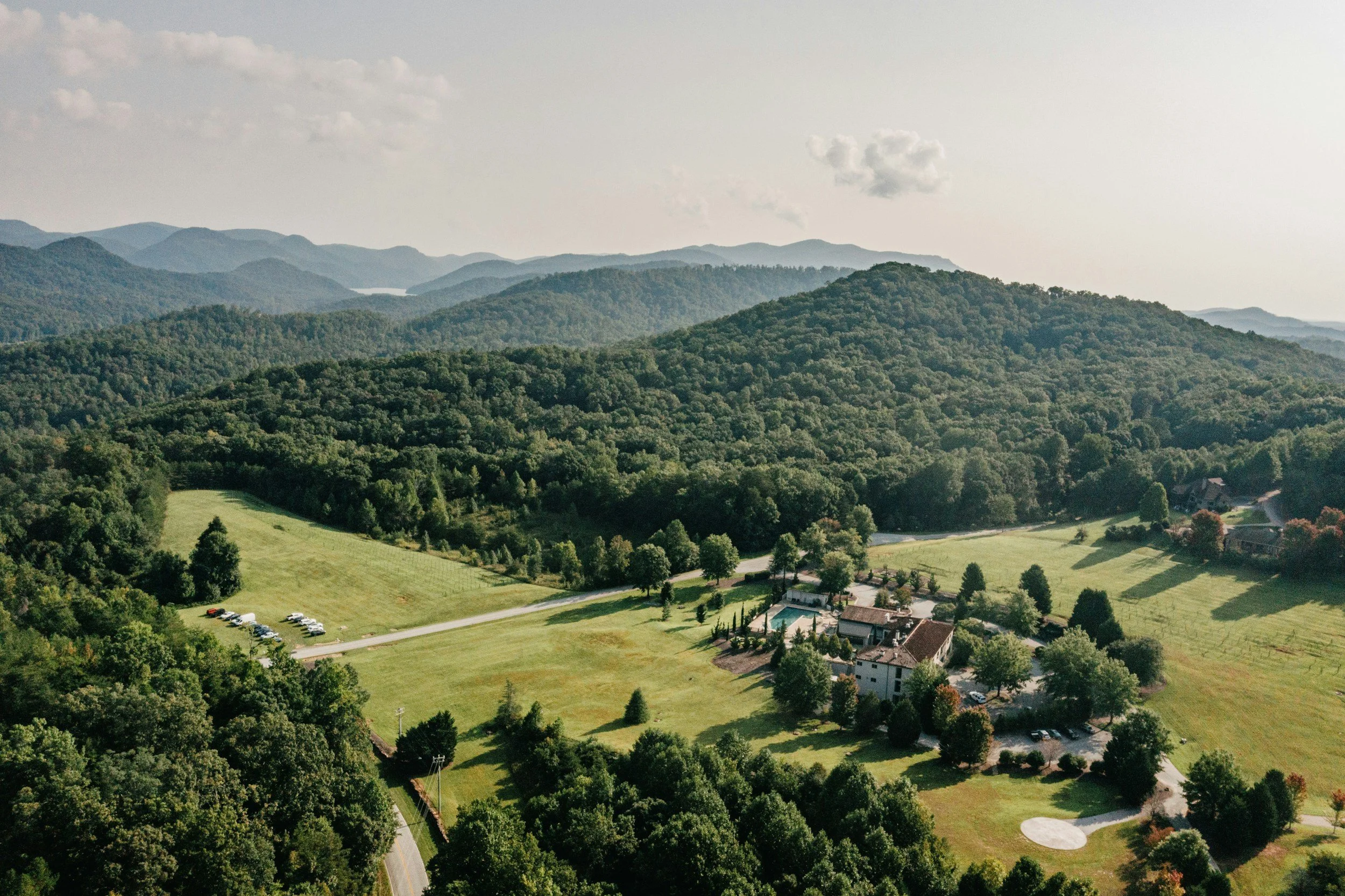 Aerial view of a mountainous landscape with lush green trees, a large open grassy area, a few buildings, and parked cars, under a partly cloudy sky.