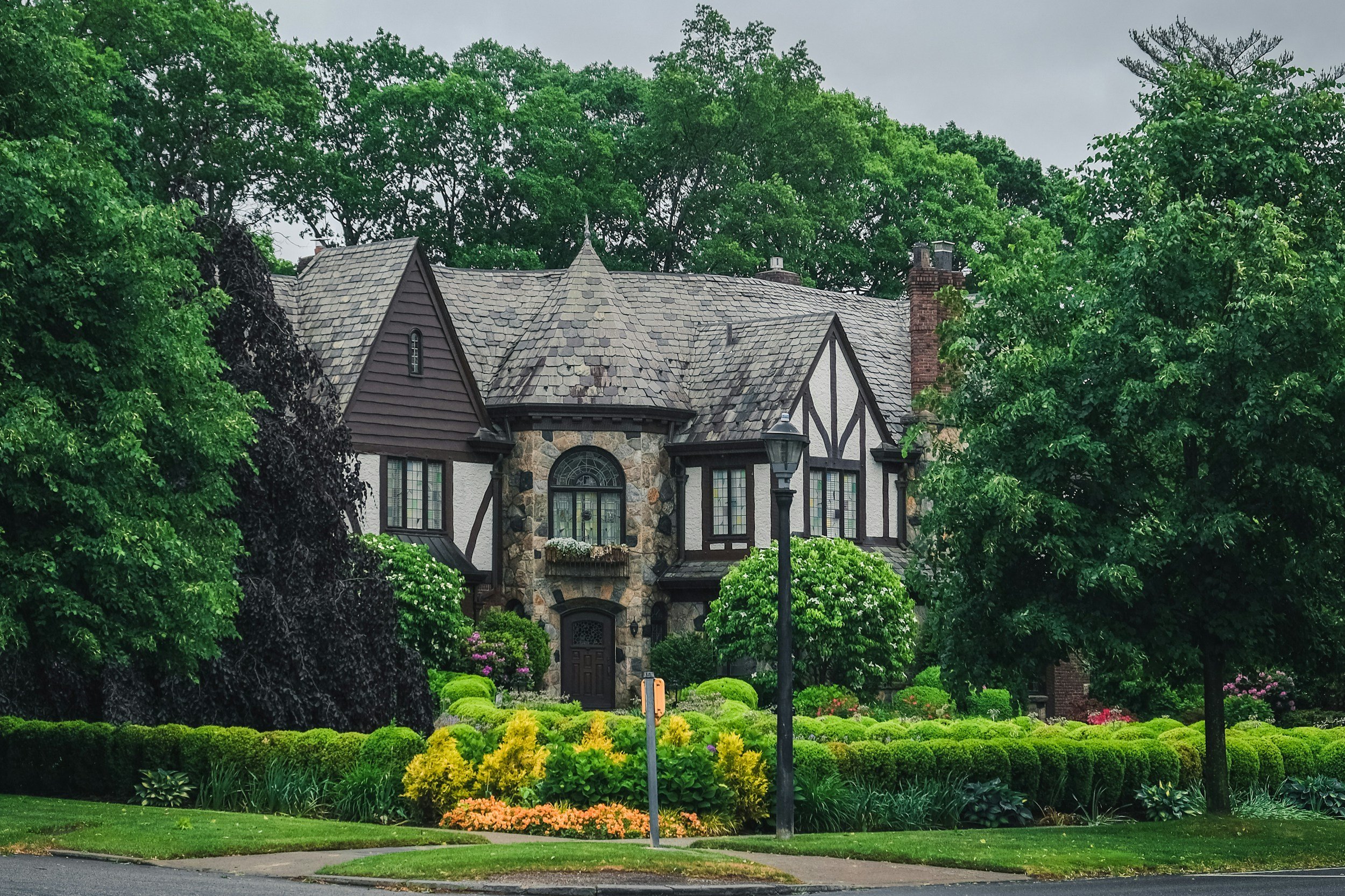 A large, Tudor-style house with a stone and half-timbered facade, surrounded by lush green trees and well-manicured garden with bushes and colorful flowers, and a streetlamp in the front.