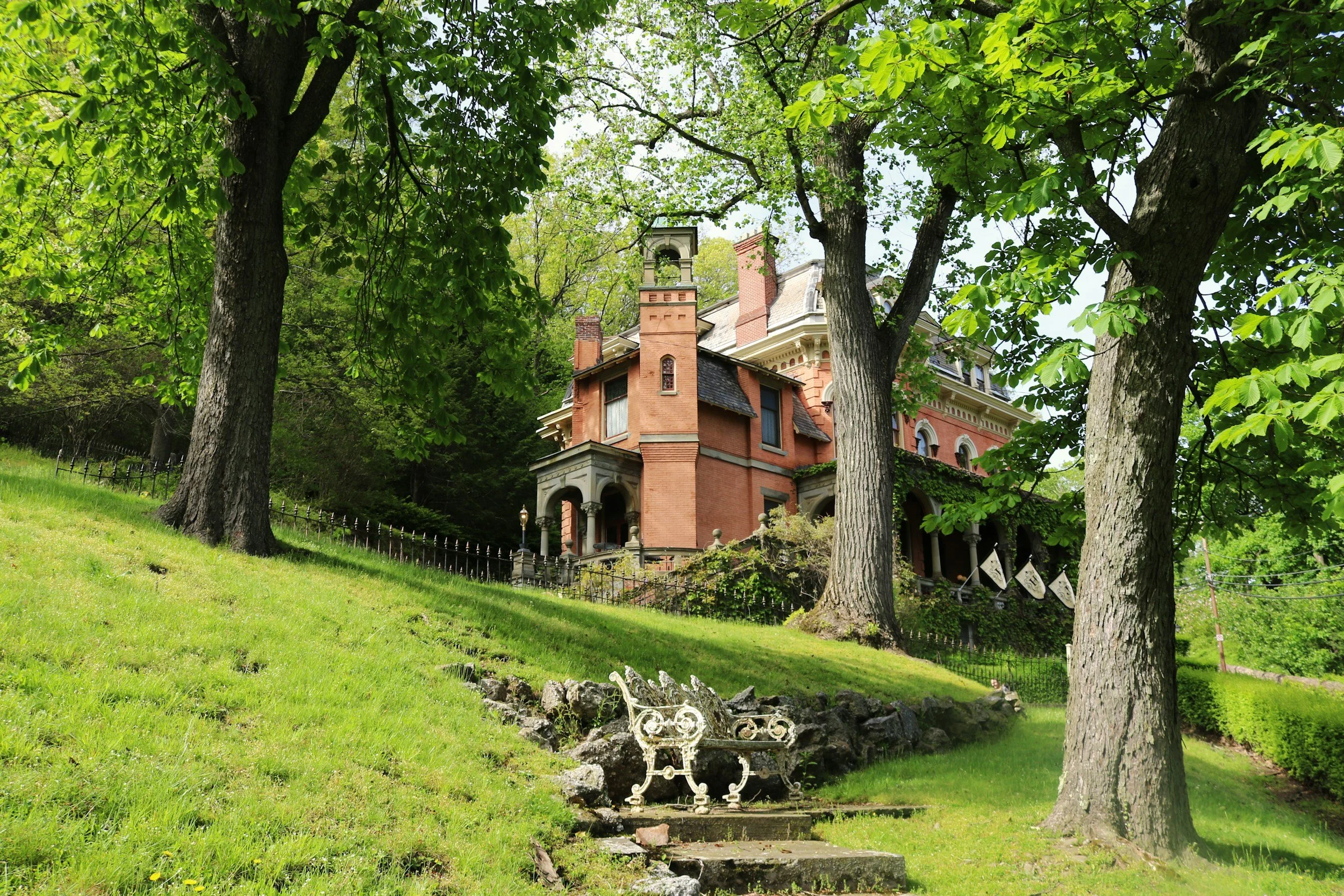 A large, historic red brick house on a hill, surrounded by green trees and grass, with a stone staircase and ornate white bench in the foreground.