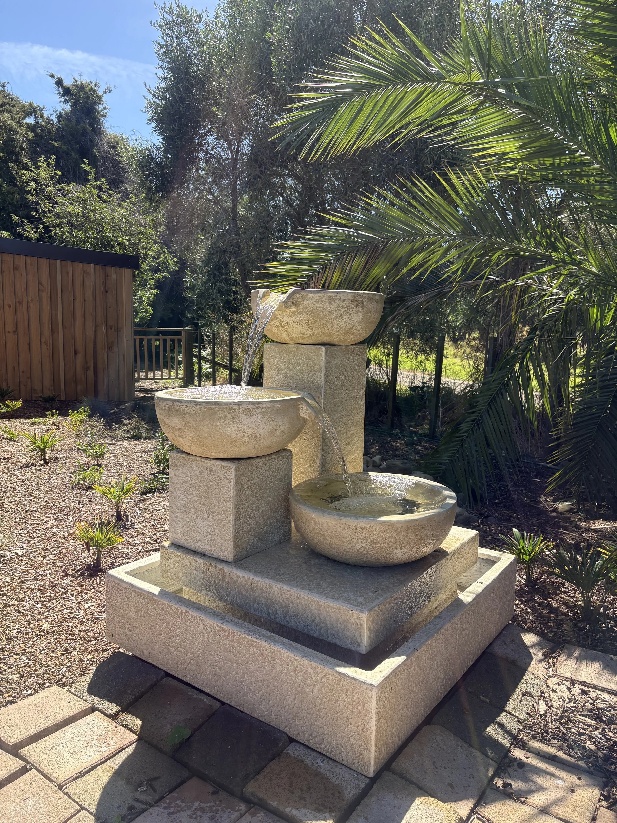 Outdoor stone fountain with three tiers, water flowing from the top to middle and then to the bottom basin, surrounded by plants and trees in a garden setting.