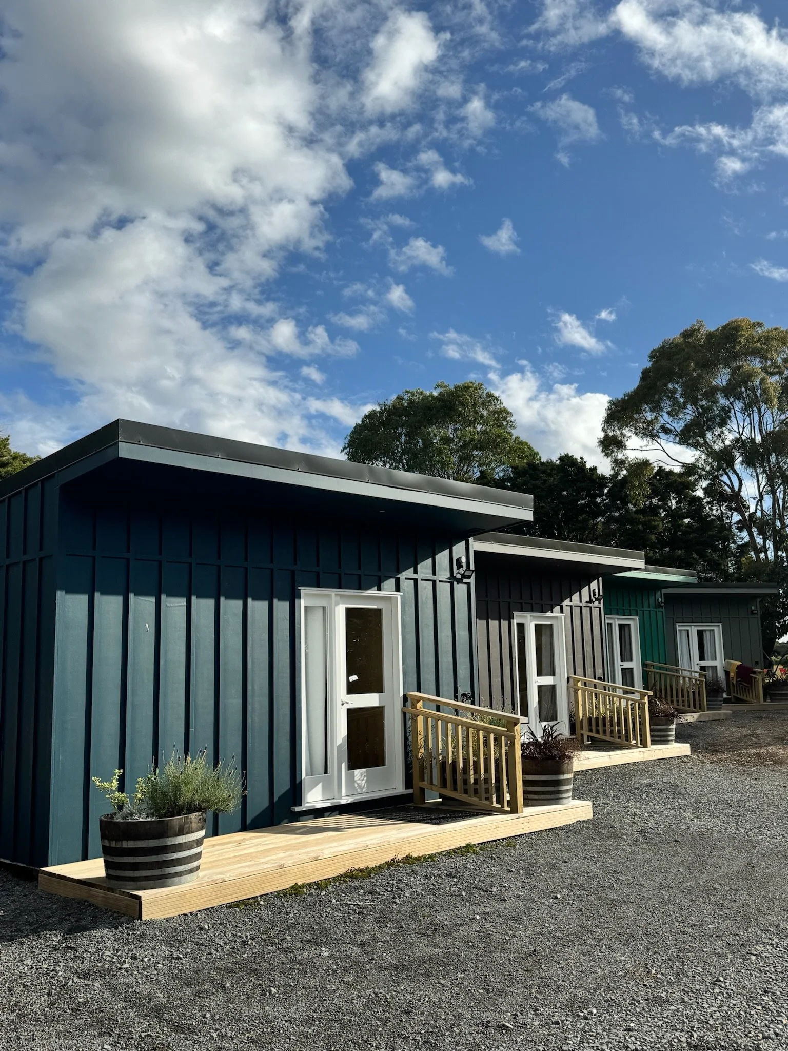 A row of small modern houses with wooden steps and potted plants, under a partly cloudy sky with trees in the background.