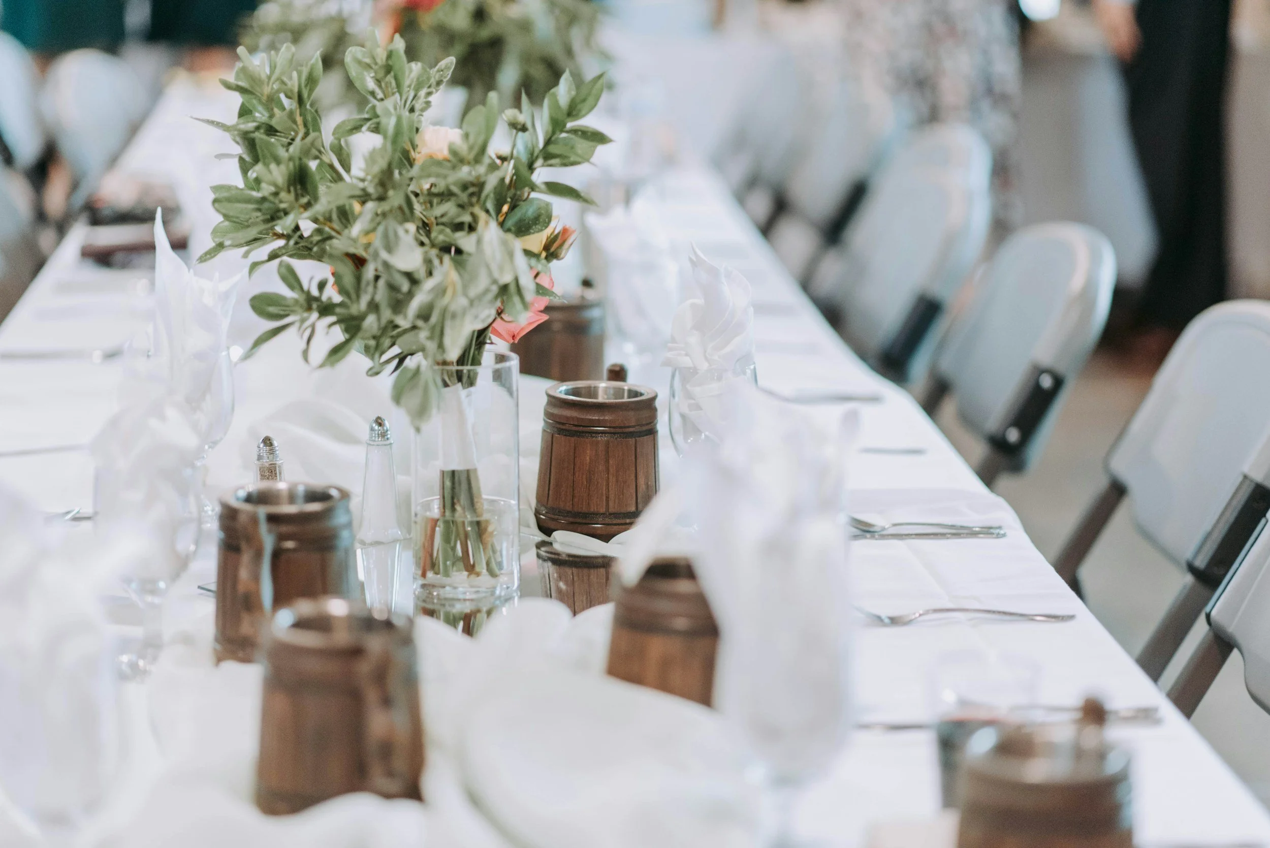 An elegantly set dining table with white tablecloth, floral centerpiece, glassware, silverware, and small wooden barrels, in a bright setting with chairs lined up along the table.