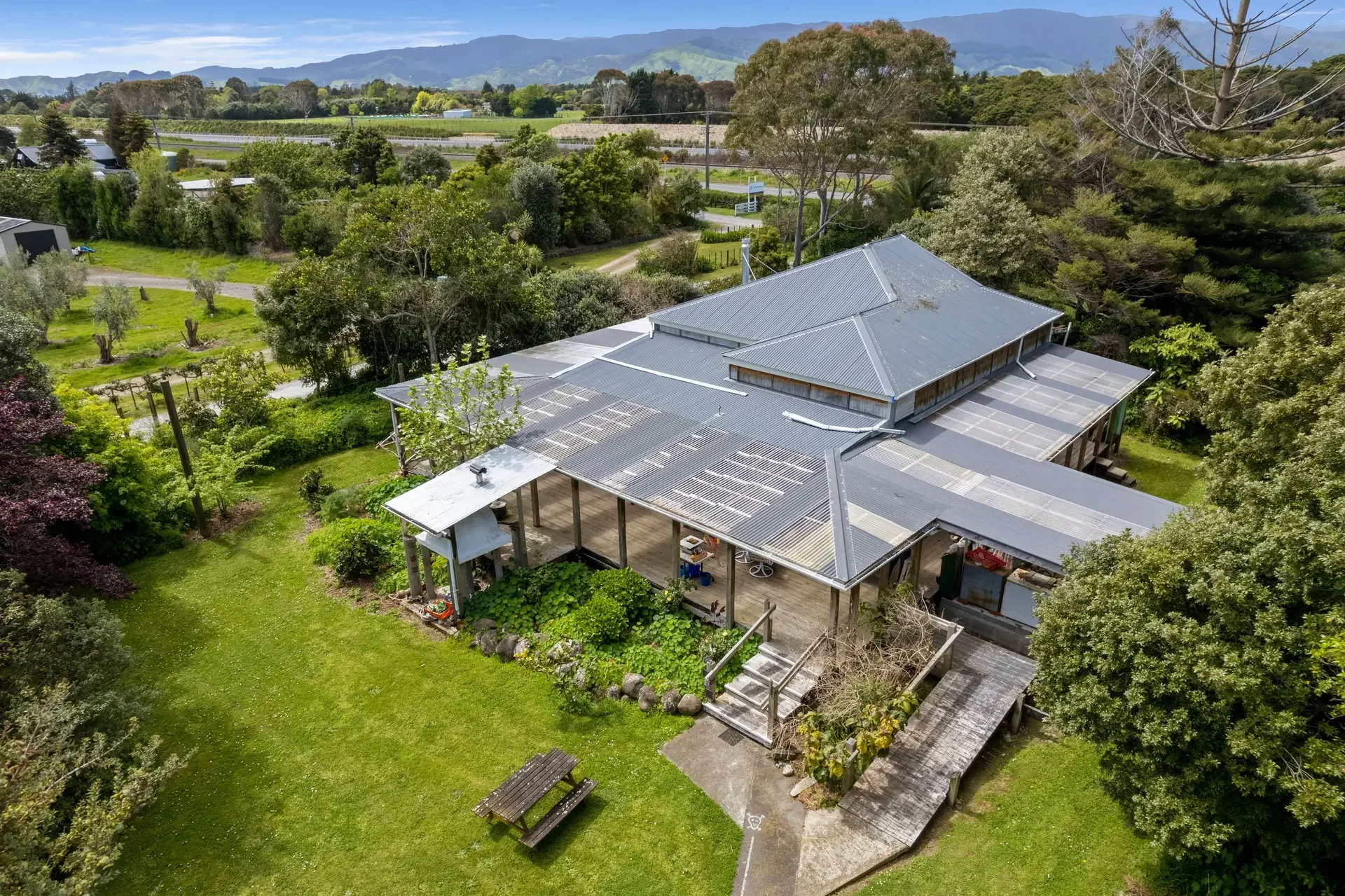 Aerial view of a large house with a metal roof, surrounded by green trees and a grassy yard with a picnic table, in a rural landscape with distant mountains and fields.