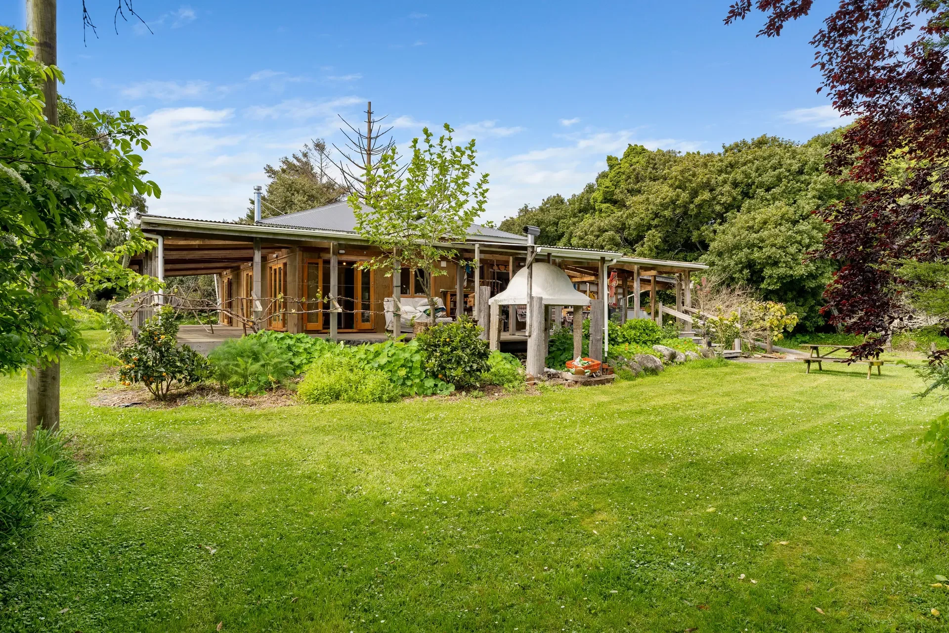 A backyard with a grassy lawn, trees, and shrubs, featuring a wooden house with a deck, potted plants, and a picnic table in the background, under a partly cloudy sky.