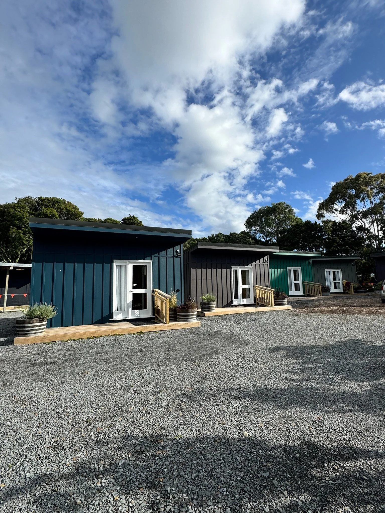 A row of small, colorful prefabricated houses with small wooden porch steps, potted plants, and gravel driveway against a backdrop of trees and a partly cloudy sky.