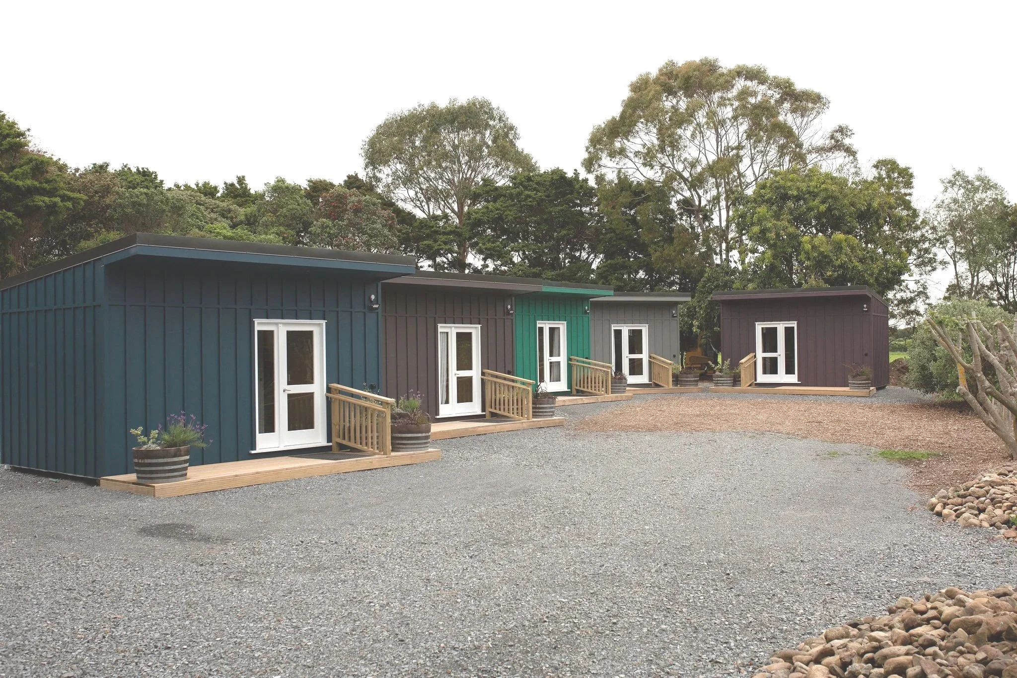 Four modern small houses with colorful exteriors in shades of blue, green, and purple, each with a white door and small wooden porch, arranged in a curve on a gravel lot with trees in the background.