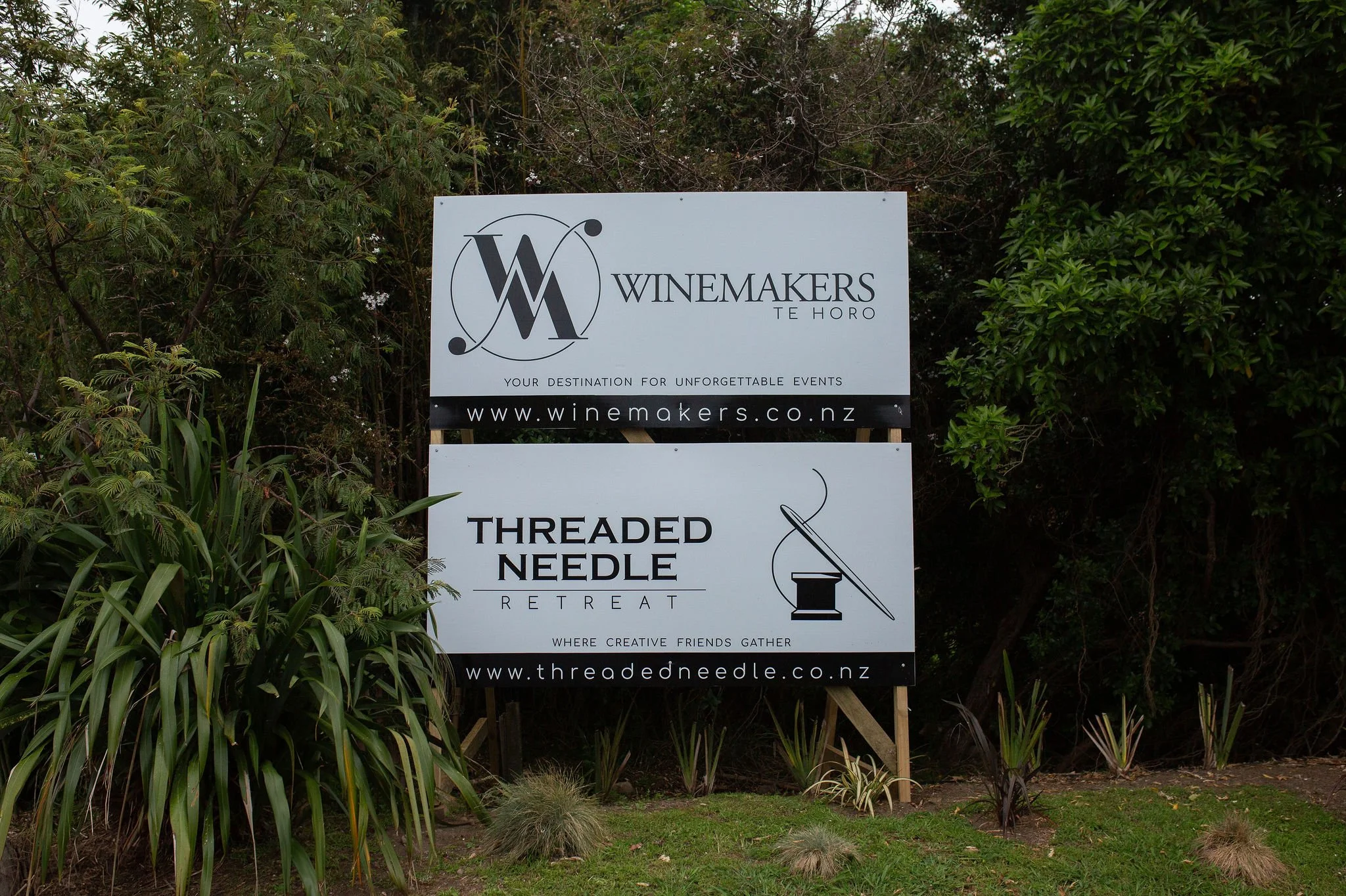 Sign for Winemakers Te Horo and Threaded Needle Retreat, featuring logos and websites, placed outdoors among greenery.