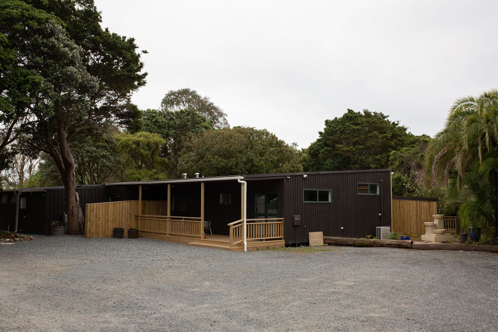 A modern, black, single-story house with a small wooden deck surrounded by trees and greenery.