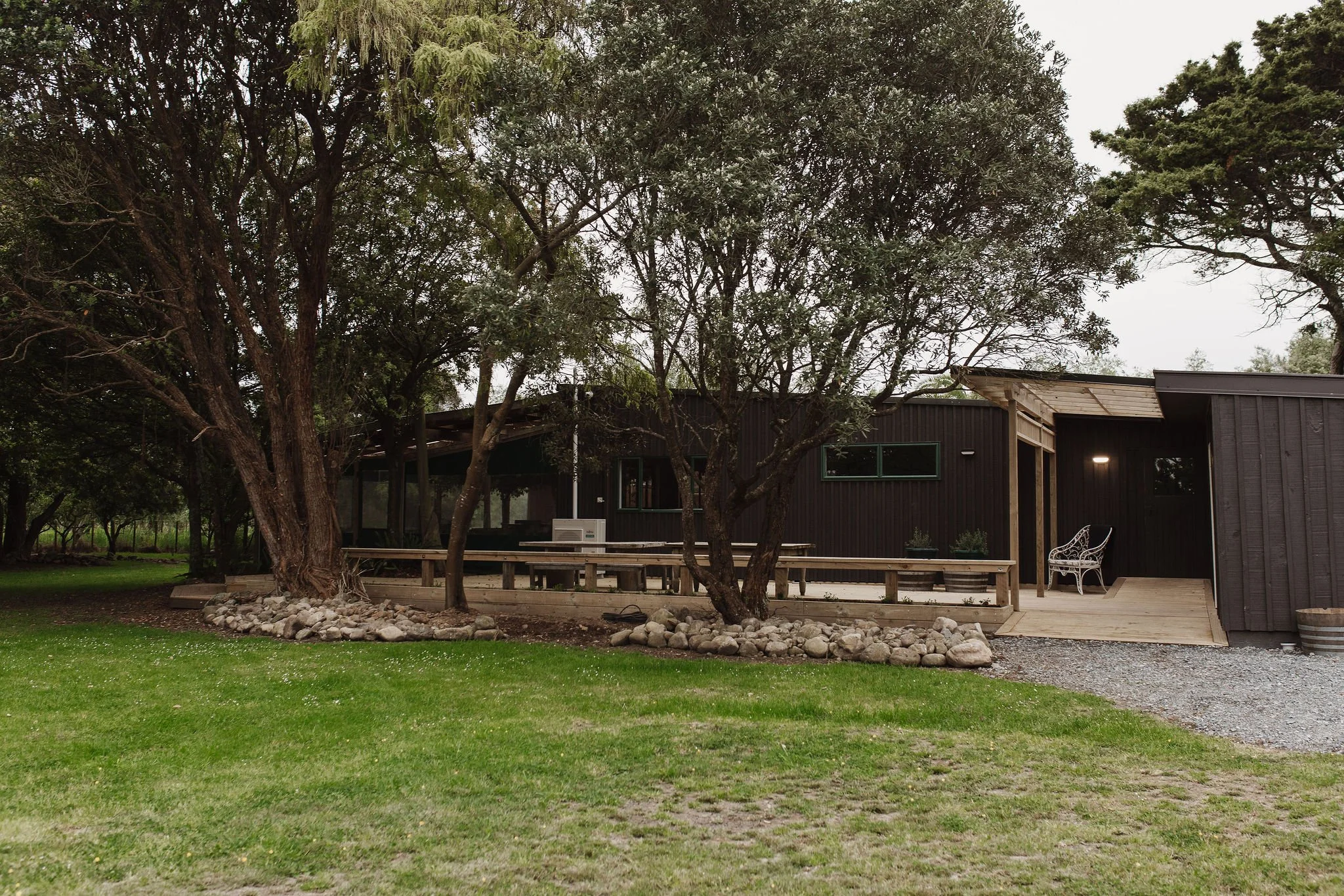A modern black house with wooden accents, surrounded by trees and a green lawn, featuring a deck with a white bench and potted plants.