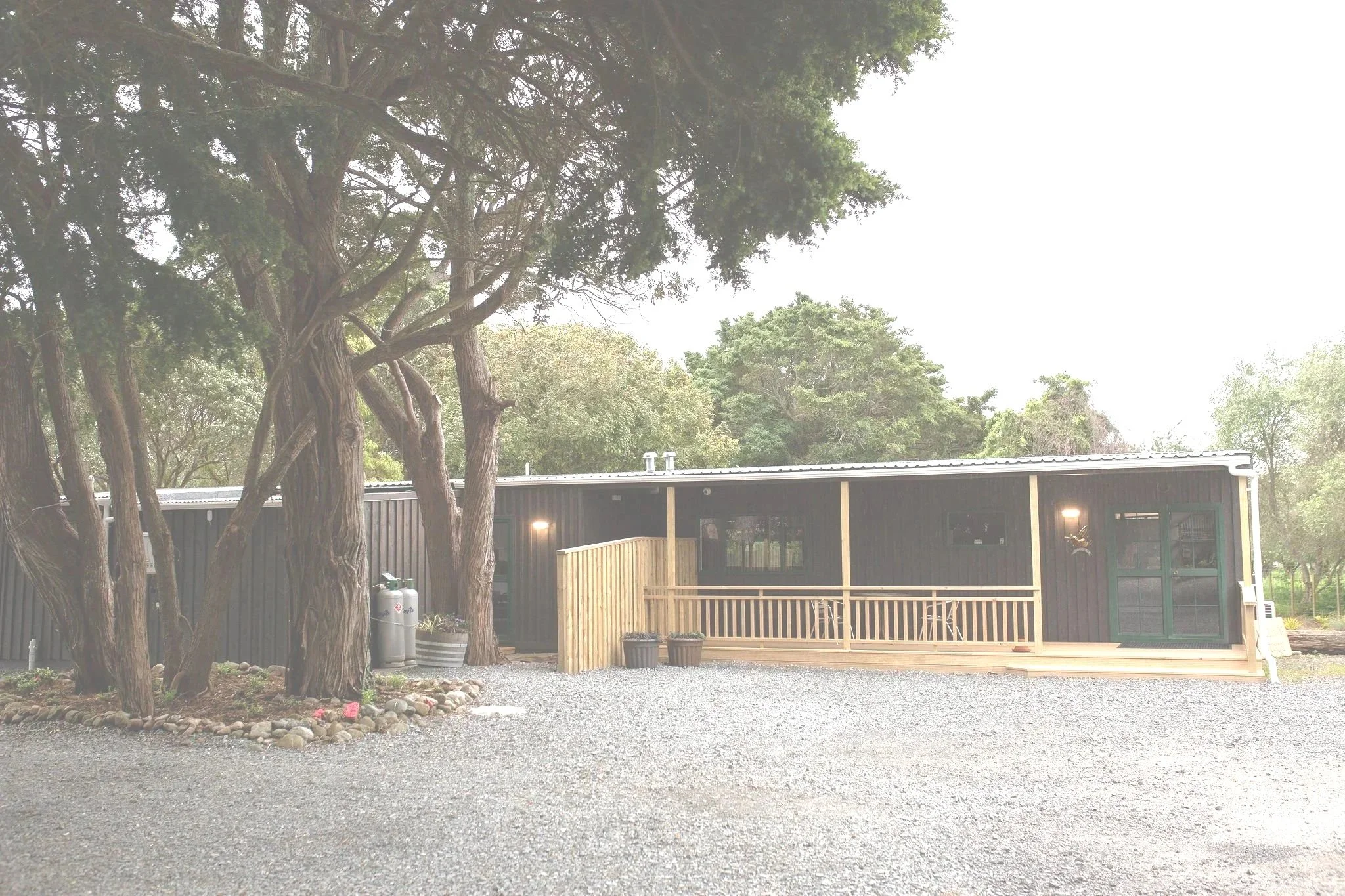 A single-story black house with a wooden porch, surrounded by trees and gravel ground.
