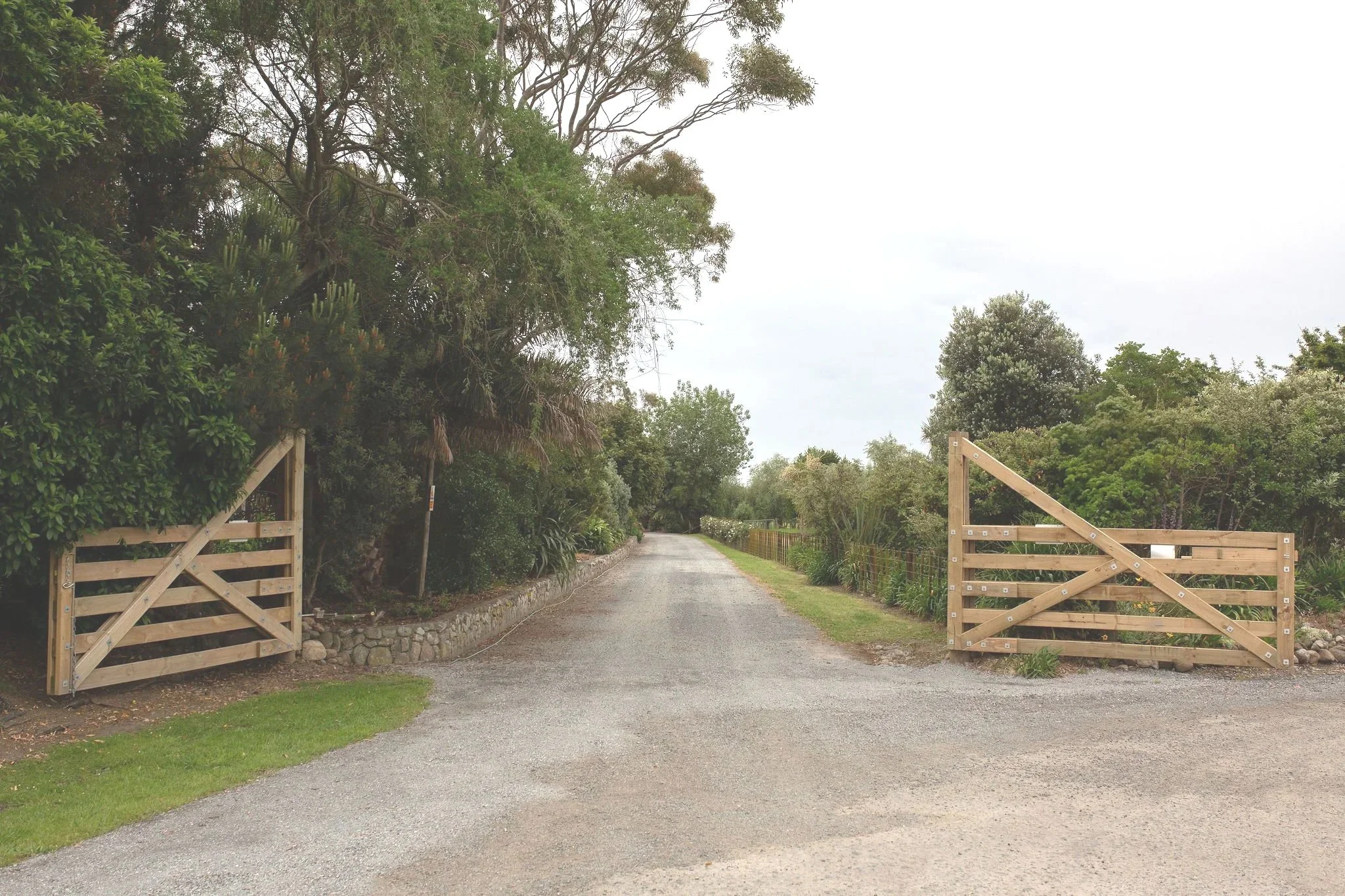 A gravel driveway or road with wooden gates on both sides, surrounded by green trees and bushes, under a cloudy sky.