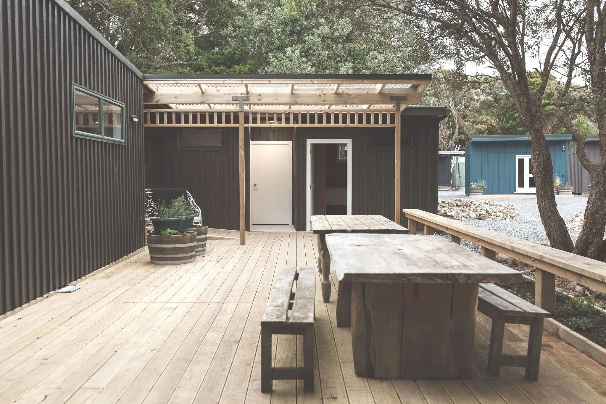 Outdoor patio area with a wooden deck, black building with doors and small window, outdoor furniture including a rustic wooden table and benches, large planters, and trees in the background.