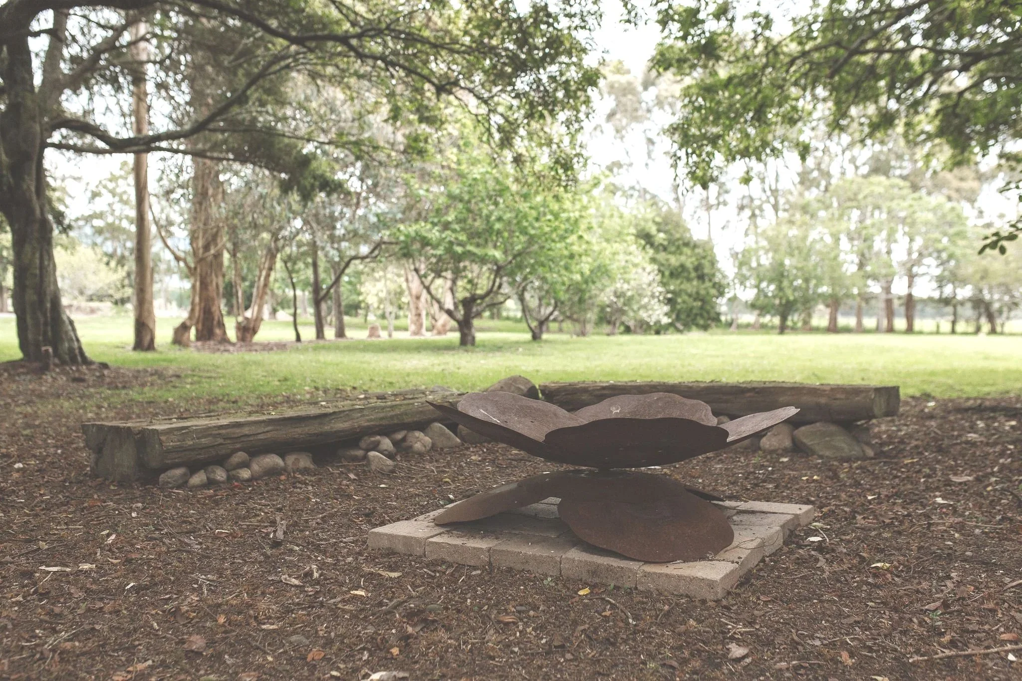 A park scene with green grass, trees, and a pile of flat stones arranged on a base of bricks, with a low wooden log in the background.