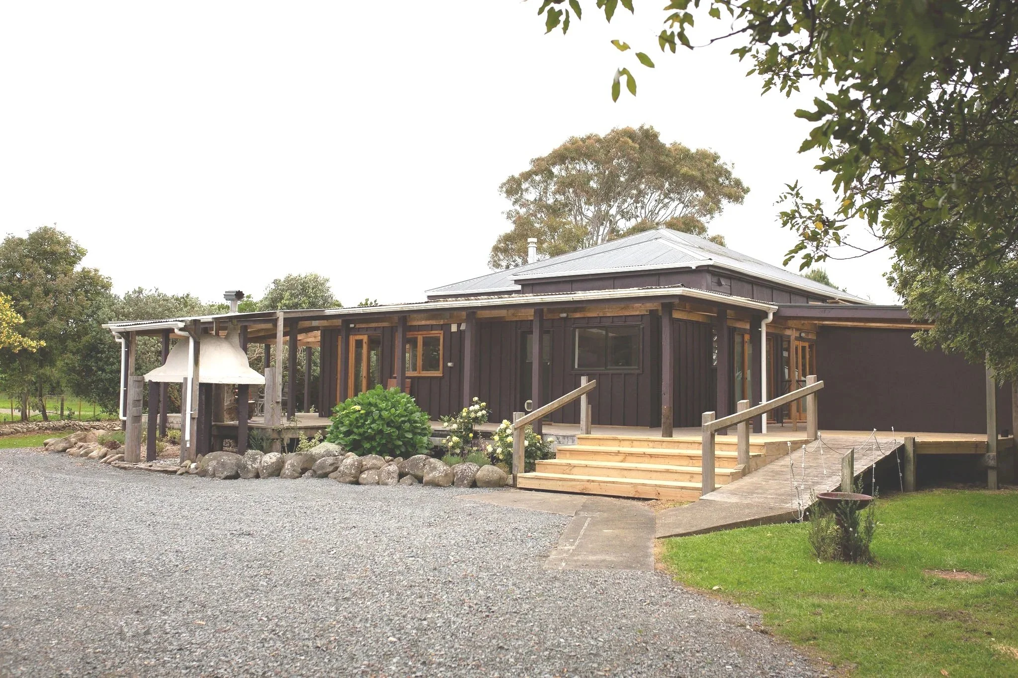 A black wooden house with a metal roof, front porch with wooden steps, surrounded by trees, gravel driveway, and a landscaped yard with stones and greenery.