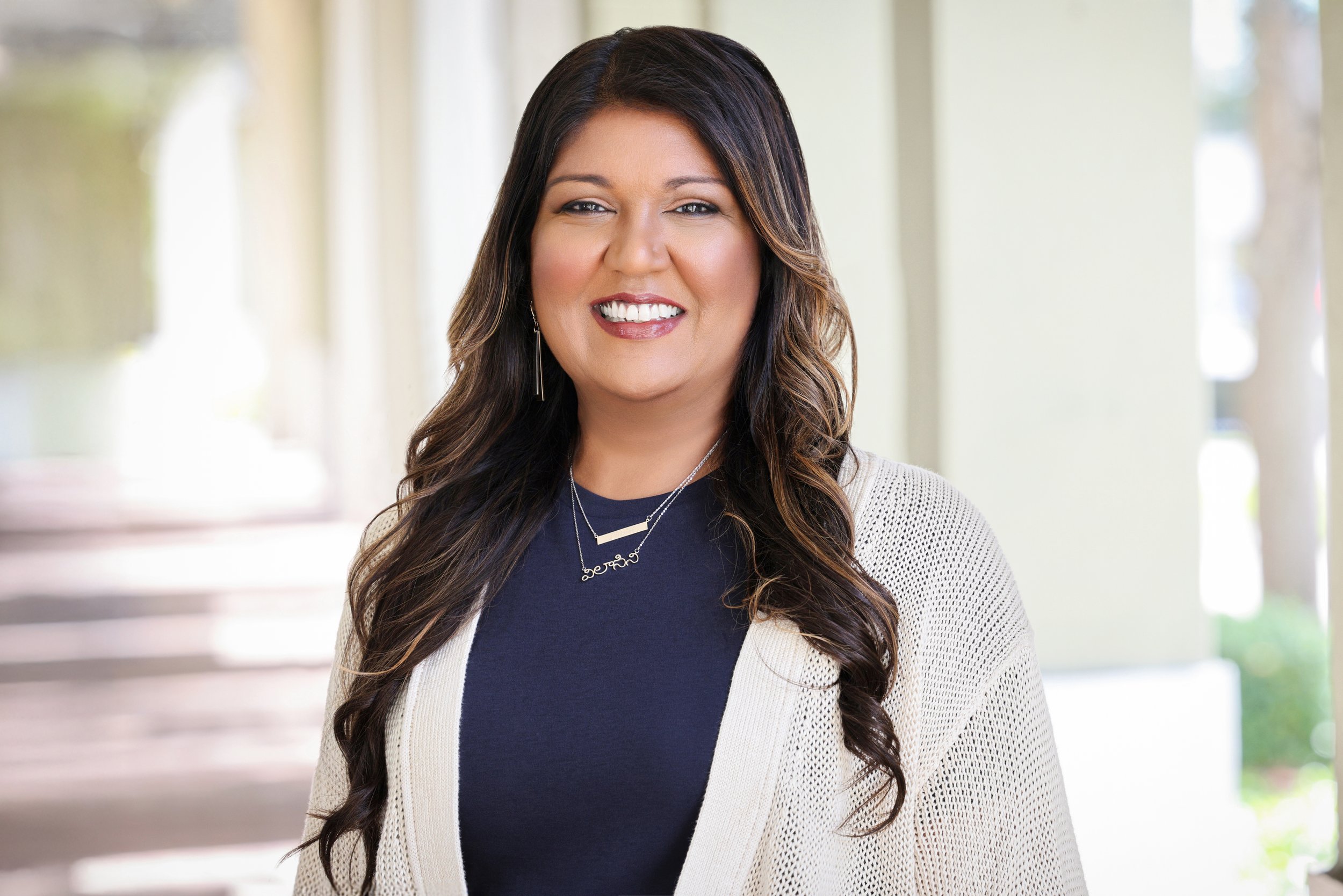 A smiling woman with long dark hair, wearing a navy blue top, a cream cardigan, and jewelry, standing outdoors with blurred background.