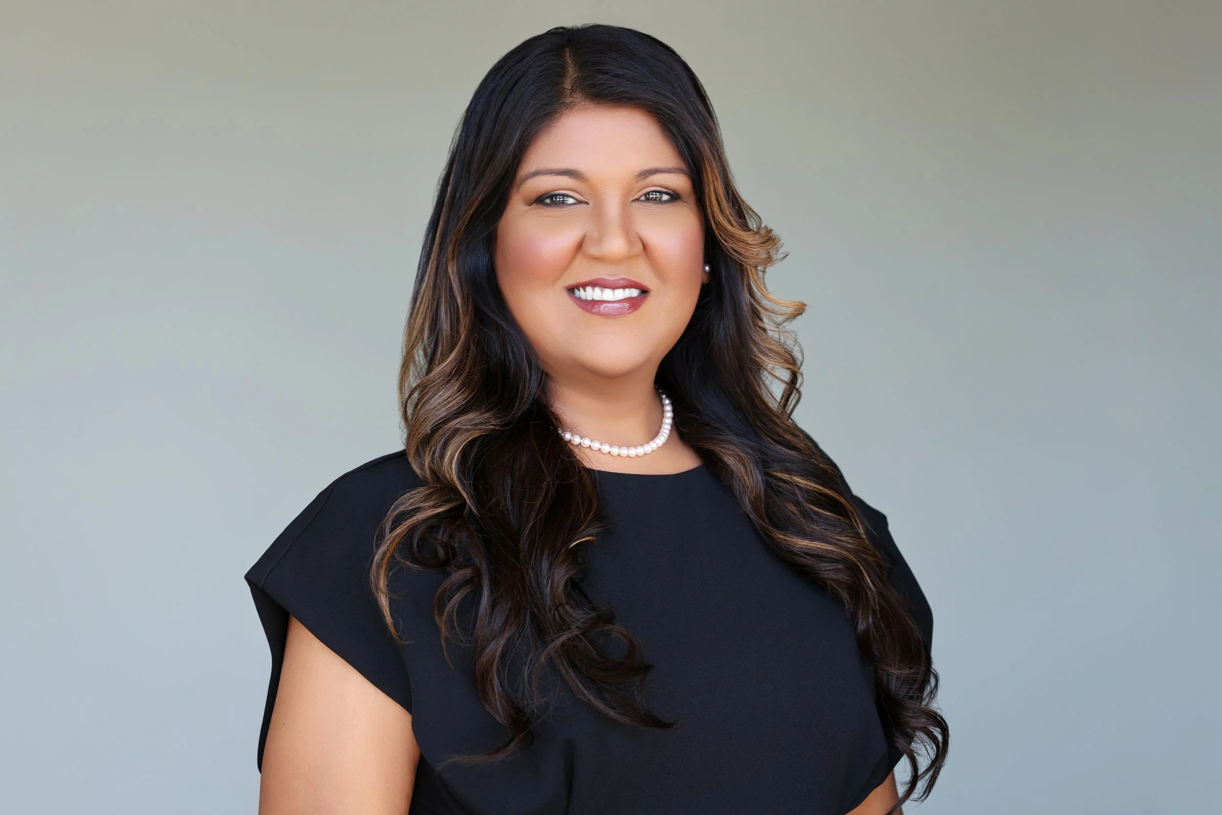A professional woman with long dark hair and pearl necklace, smiling in a black top, with a neutral blurred background.
