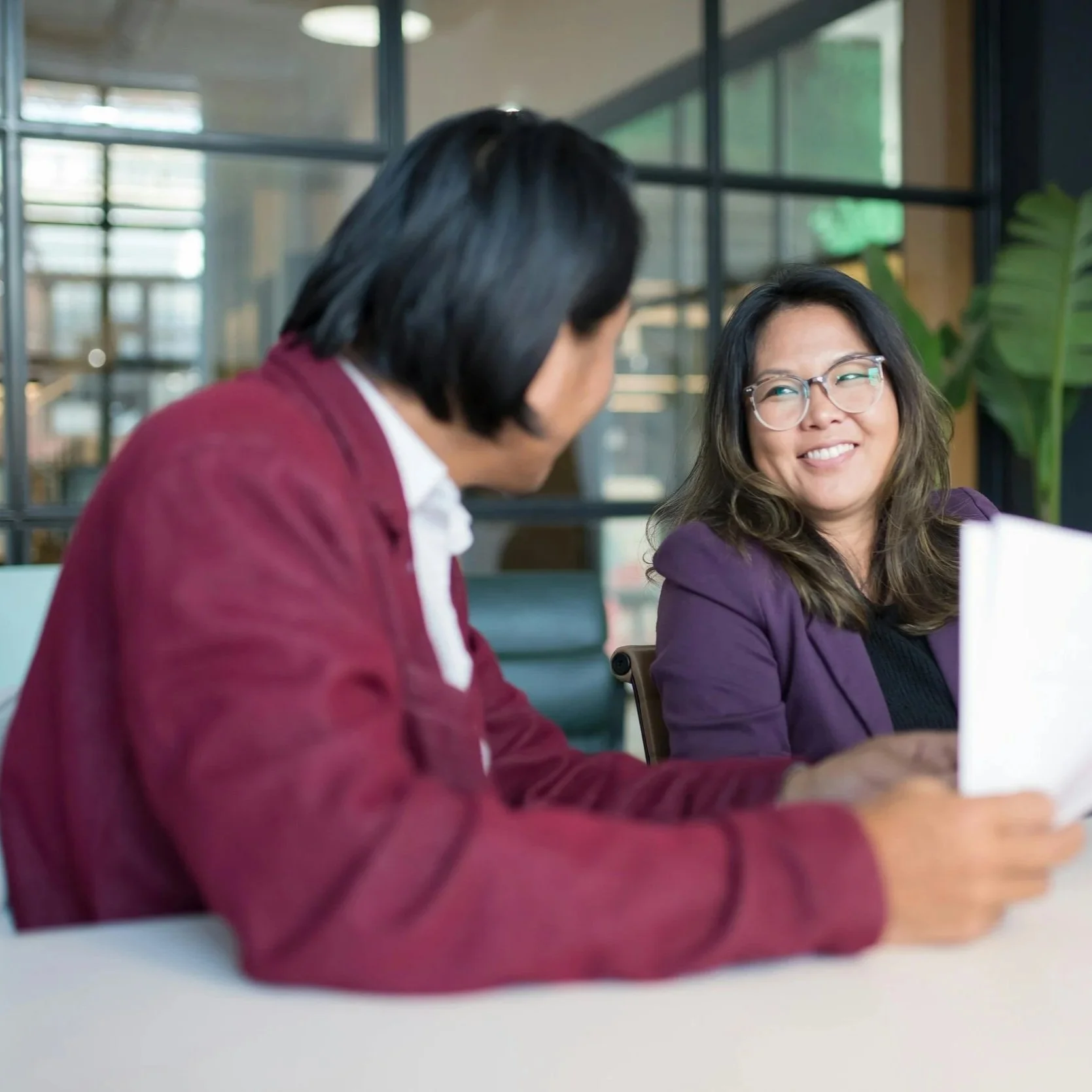 Two women sitting at a table, engaged in conversation, with one holding a notebook and smiling at each other in a modern office setting.