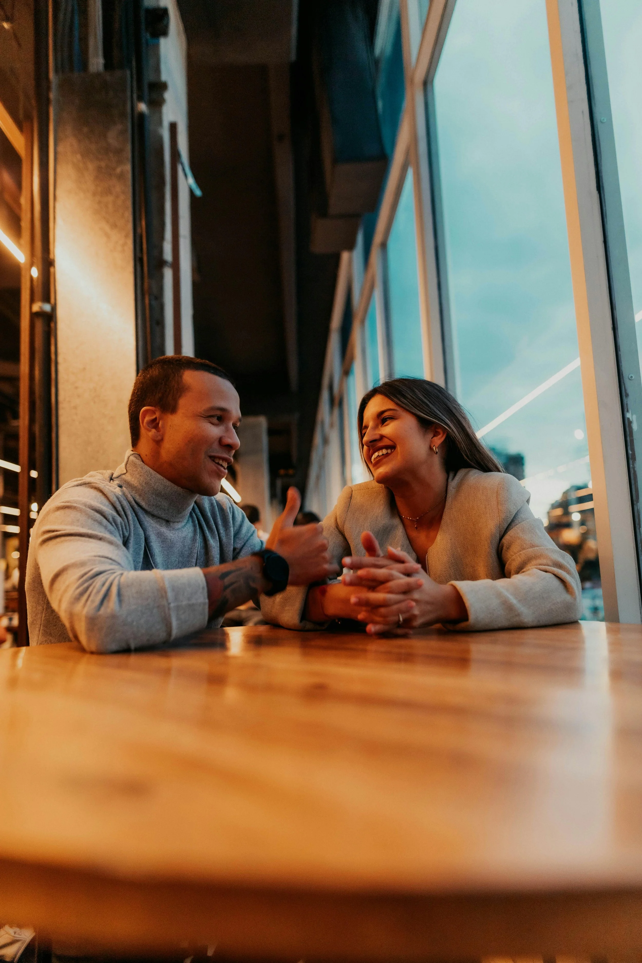 A man and woman sitting at a table in a cafe, smiling and talking, with large windows showing an outdoor scene in the background.