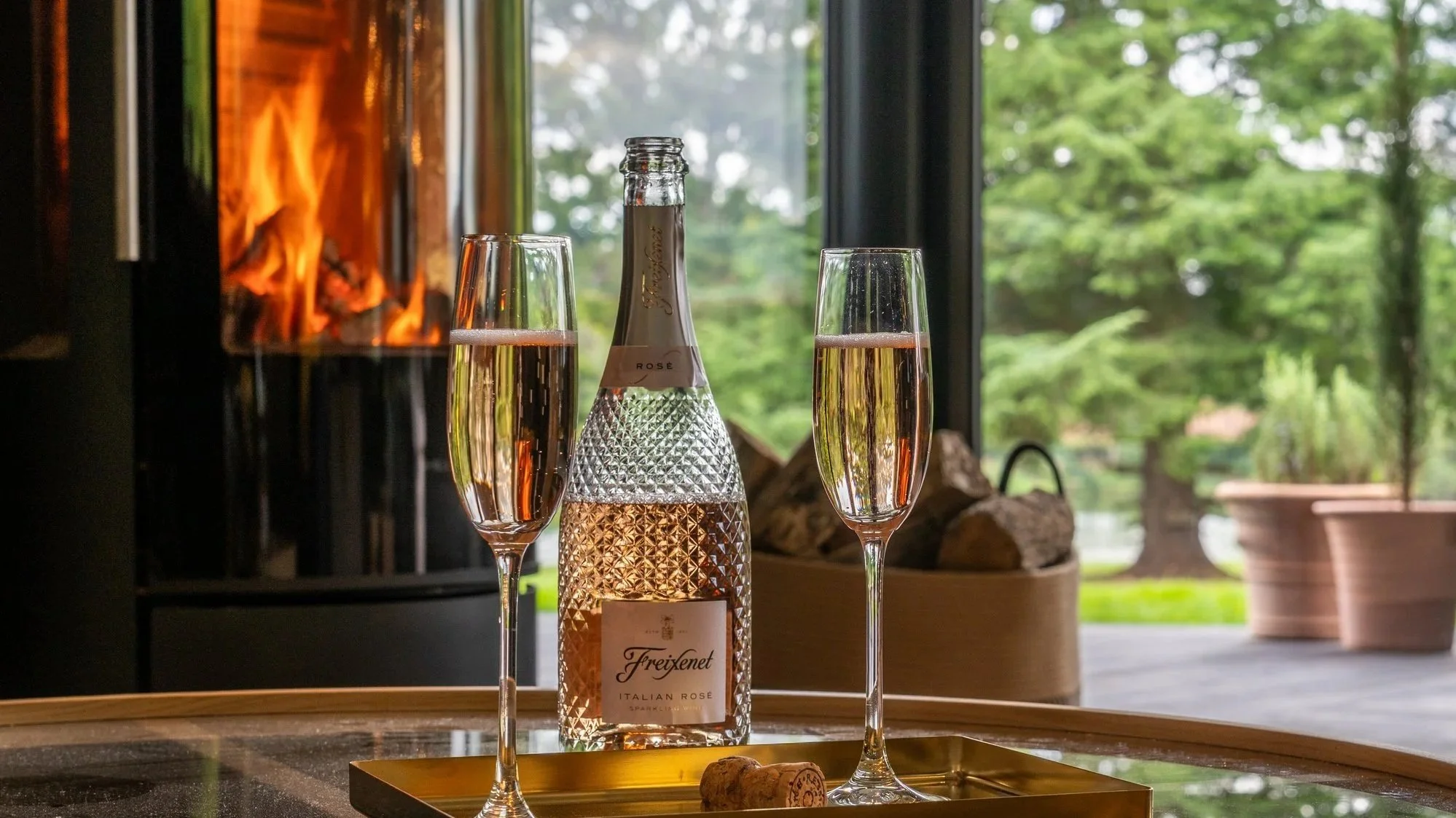 A bottle of Freixenet Italian Rosé sparkling wine with two filled glasses on a gold tray, in front of a modern fireplace with a fire burning, and a large window showing green trees outside.
