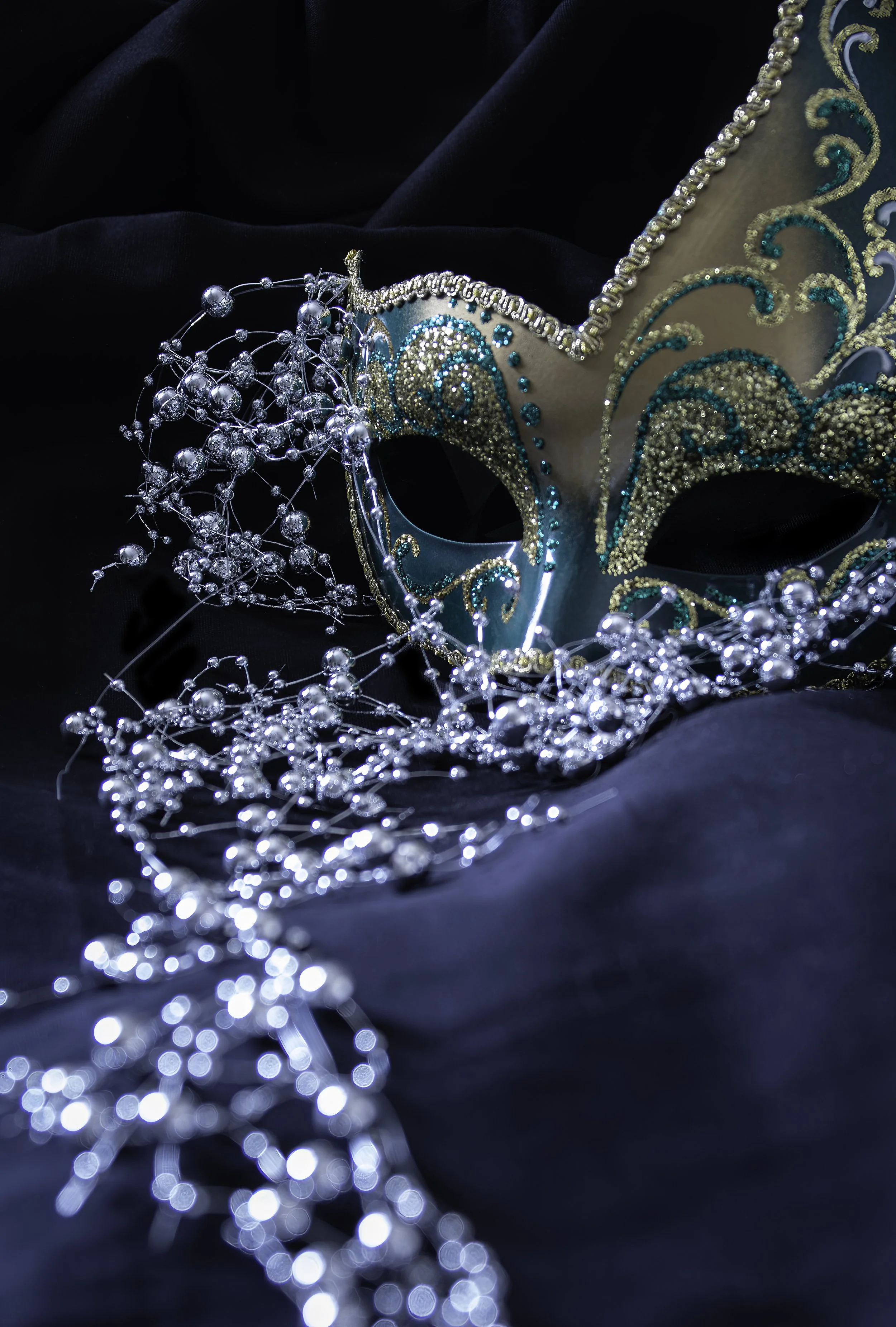A decorative Venetian-style mask with teal and gold accents, embellished with beads and intricate gold swirl patterns, surrounded by a silver beaded necklace on black fabric.