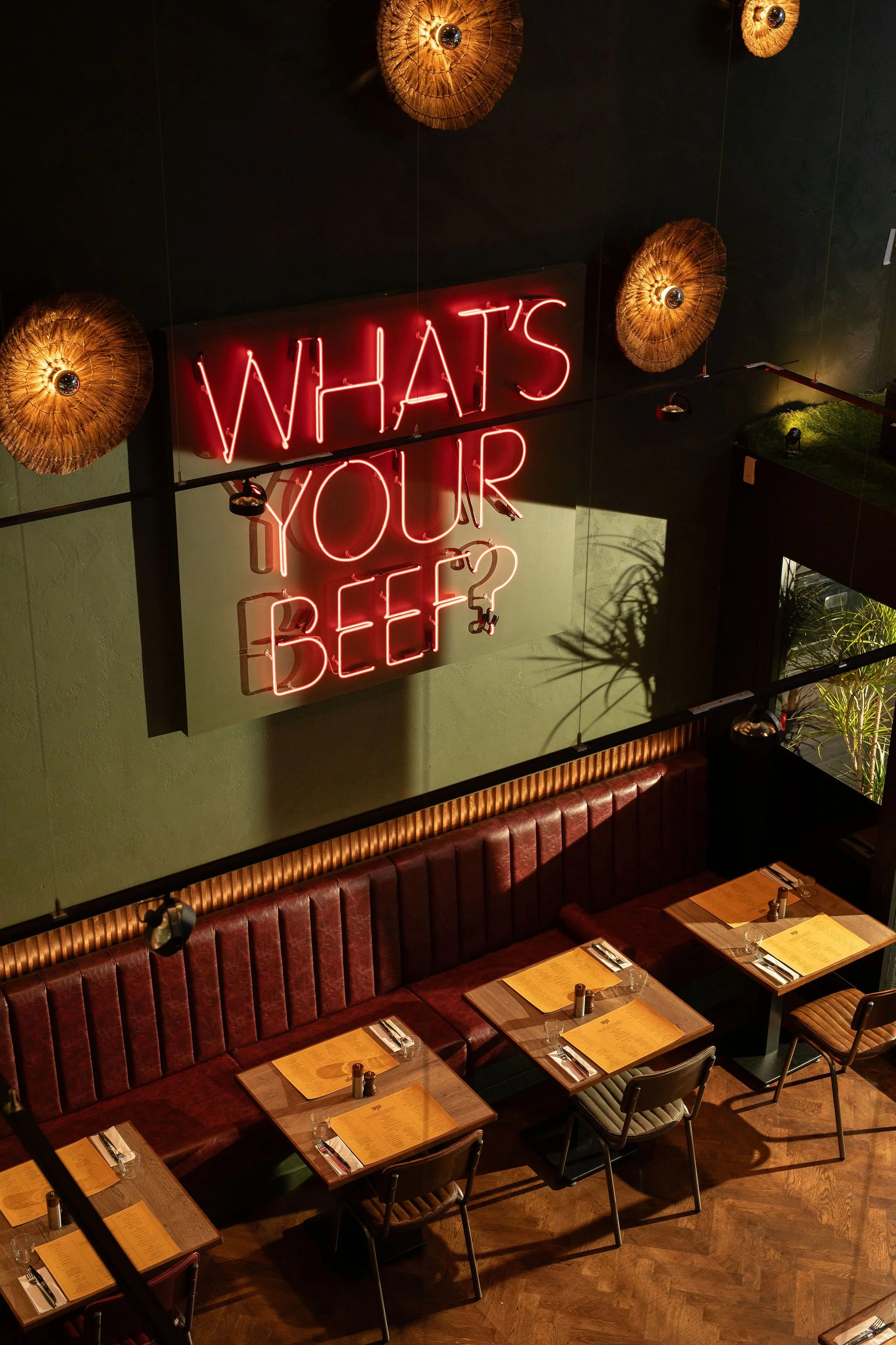 Interior view of a restaurant with a neon sign reading 'What's your beef?' on a green wall, surrounded by decorative woven light fixtures, with a red leather booth and tables set with menus, utensils, and glasses.