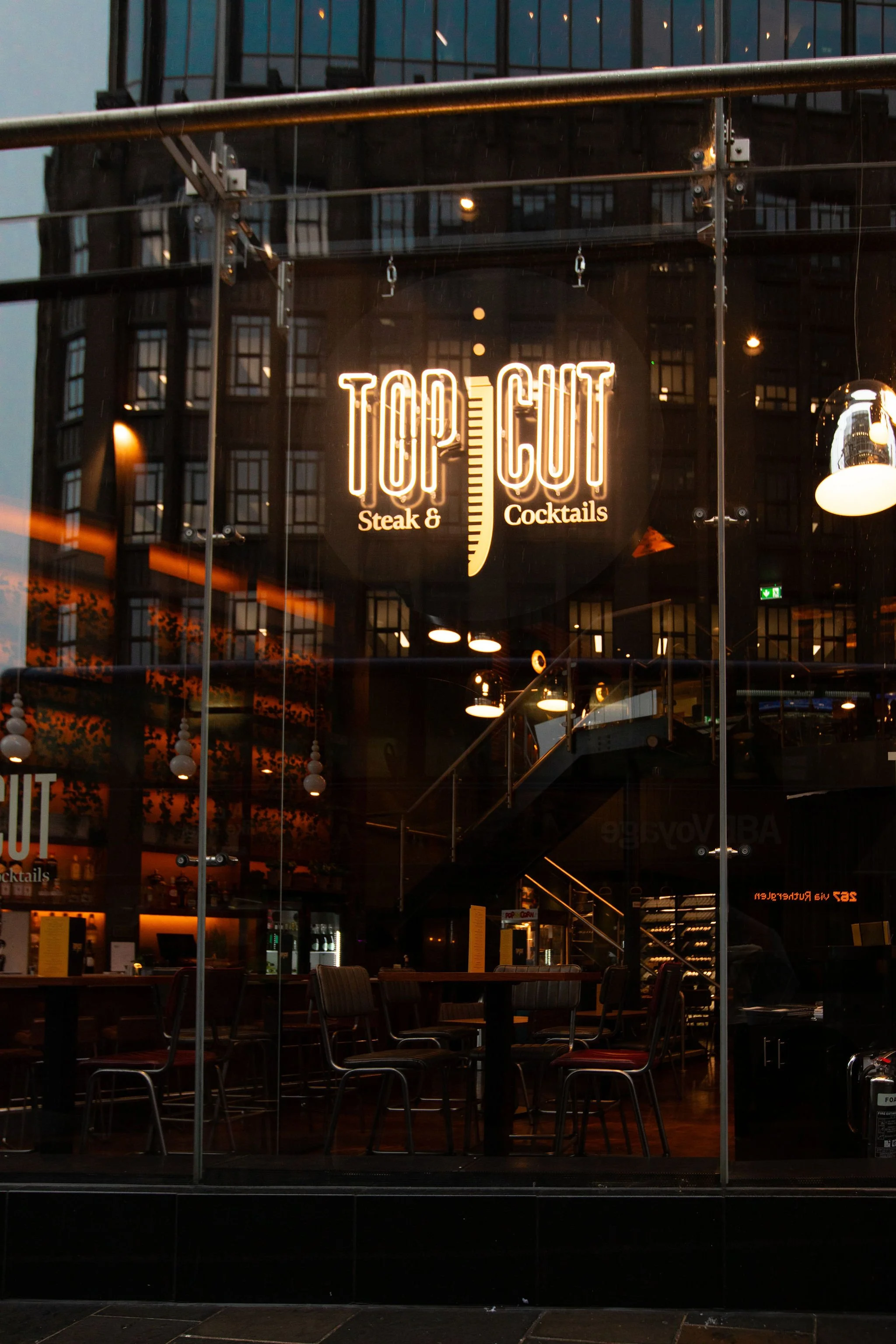 Neon sign reading "Top Cut Steak & Cocktails" inside a restaurant, viewed through a glass window, with visible tables and chairs.