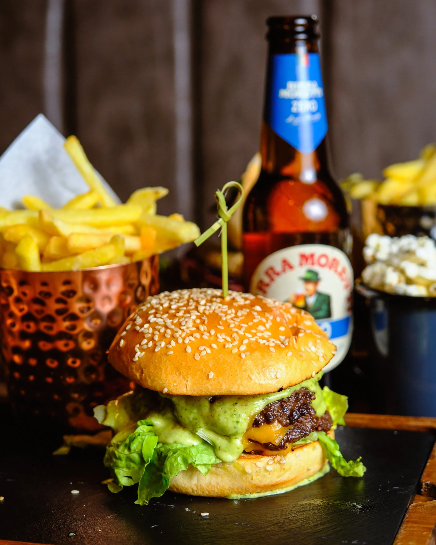A cheeseburger with lettuce, cheese, and sauce in a sesame seed bun, served with French fries, a bottle of beer, and sides of potato salad and coleslaw.