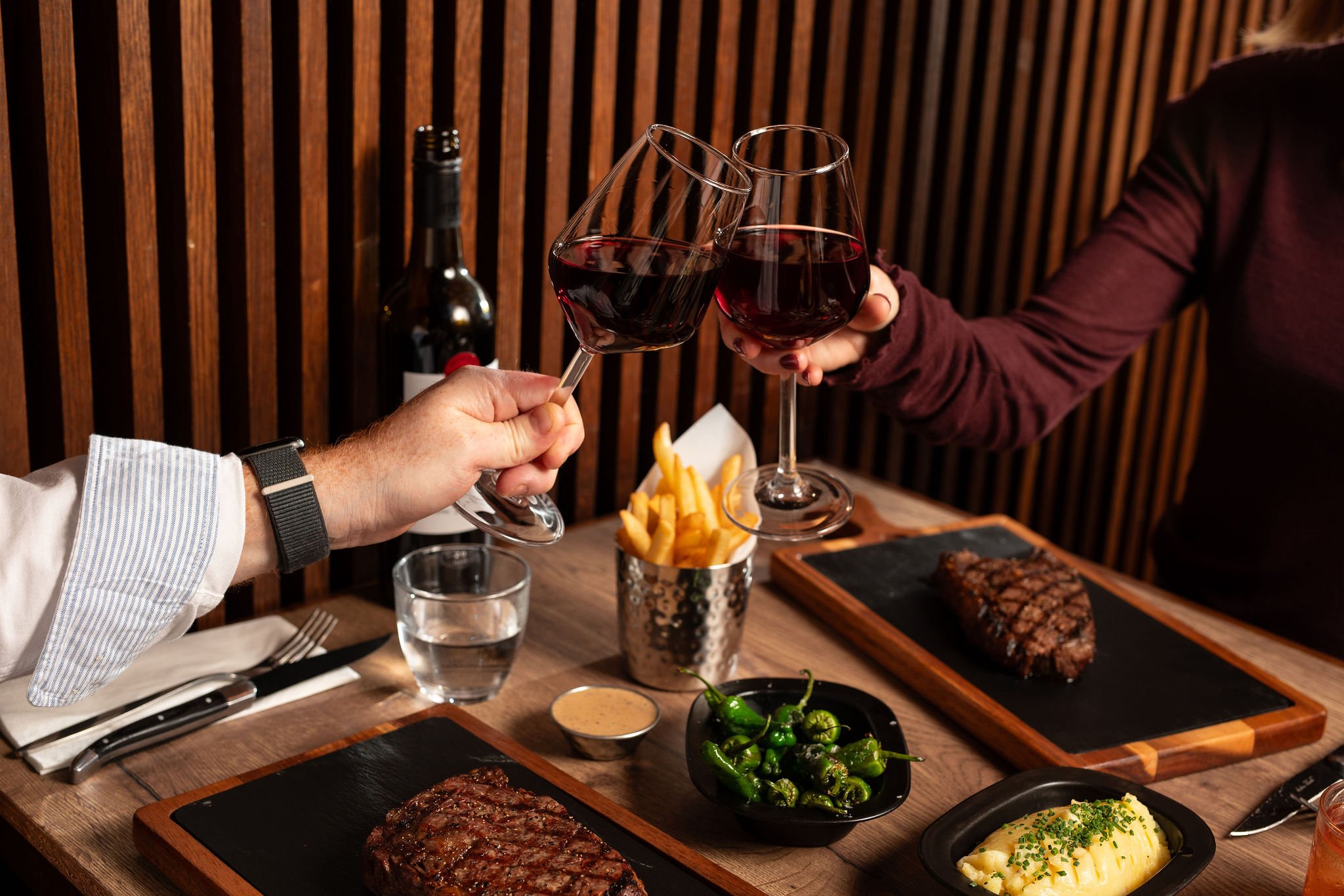 Two people clinking glasses of red wine at a steak dinner table with steaks, fries, green peppers, mashed potatoes, and a glass of water.