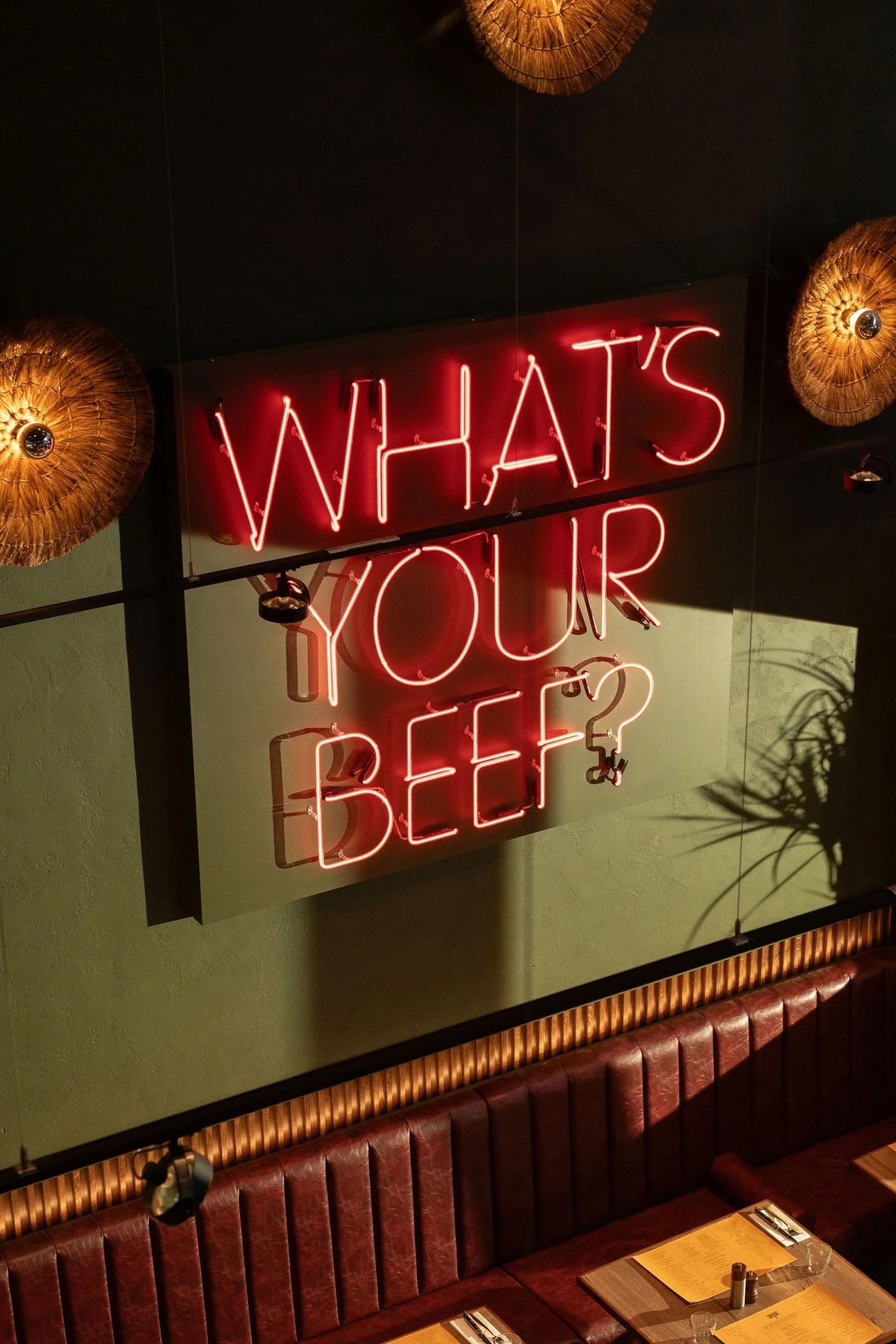 Neon sign reading 'WHAT'S YOUR BEEF?' hanging above a restaurant booth with wood paneling and table settings, decorated with wicker lamps and a shadow of a plant on the wall.