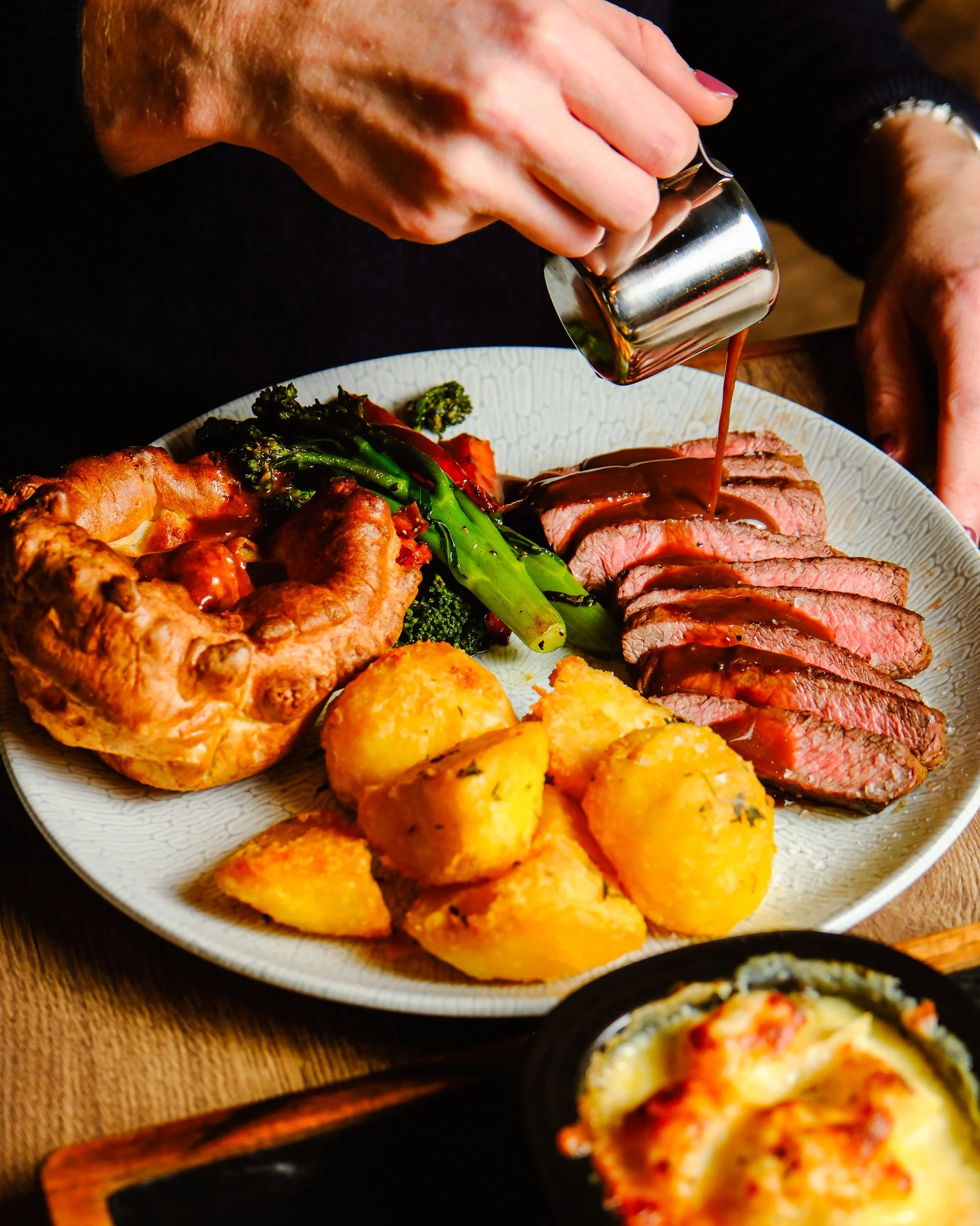 Person pouring gravy over sliced beef steak with roasted potatoes, broccoli, and a Yorkshire pudding on a dinner plate.