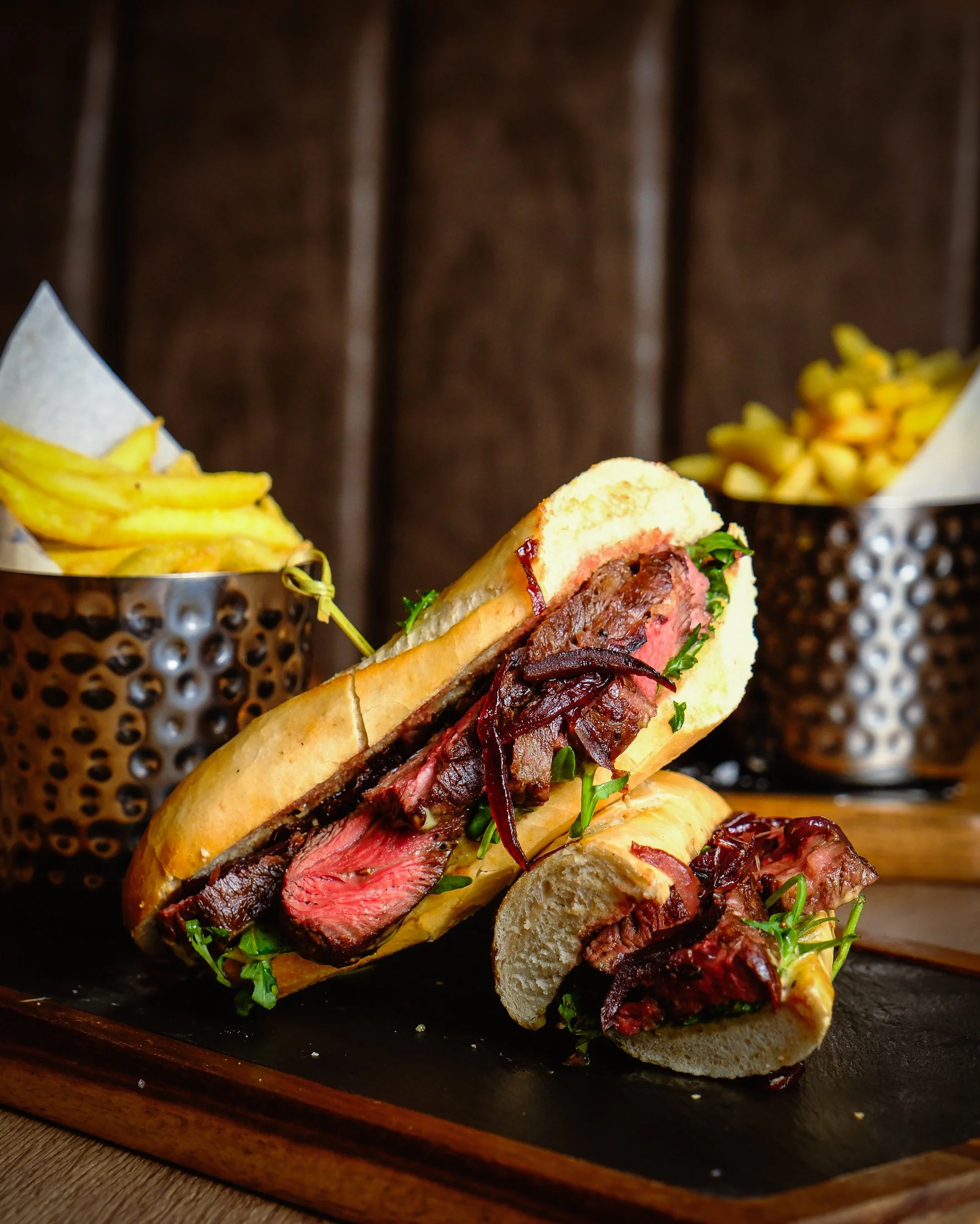 Close-up of a fork holding a piece of cooked steak topped with chopped green herbs, with more slices of steak on a black platter in the background, and a pile of french fries in the foreground.