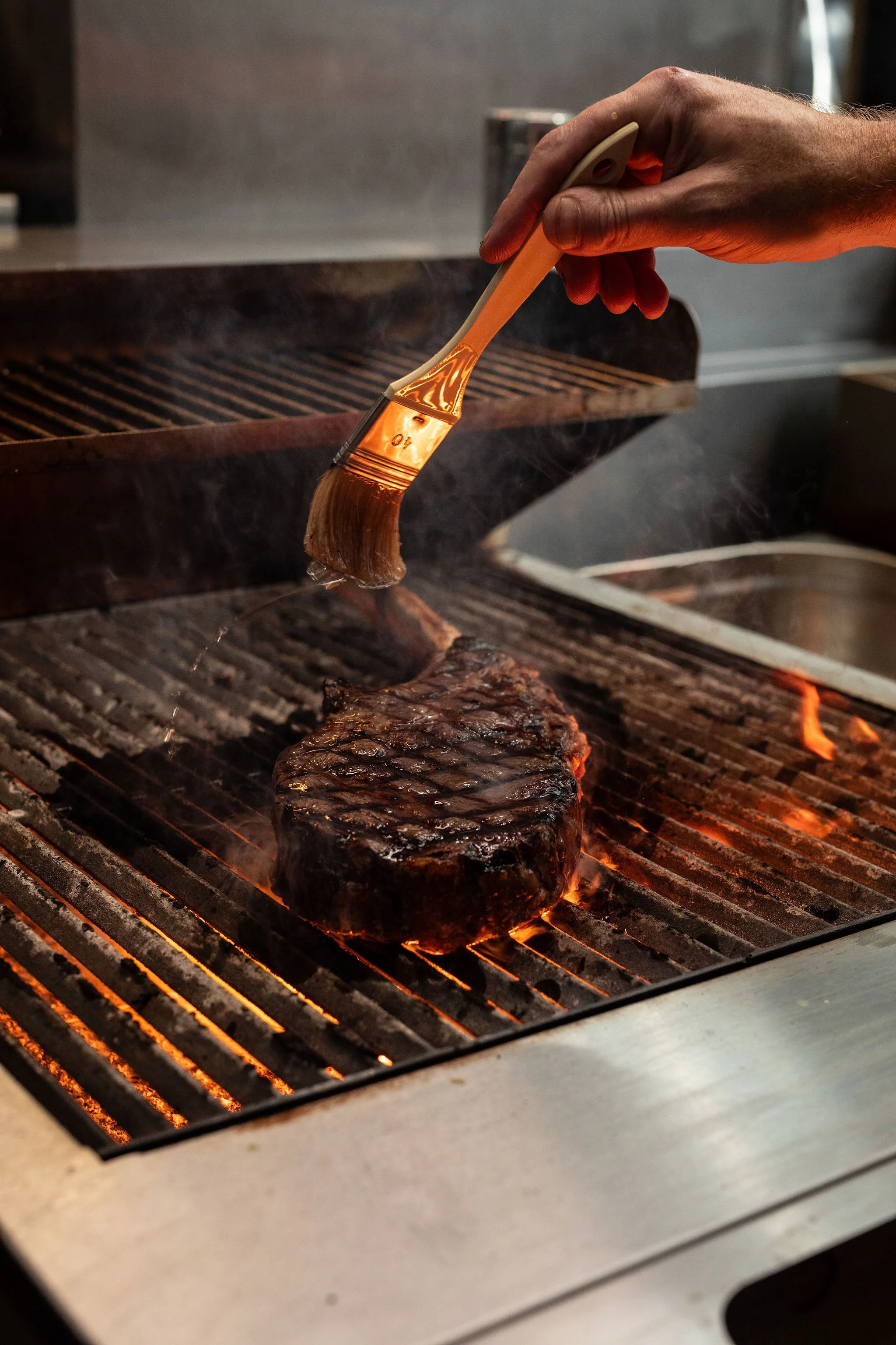 Person using a brush to apply sauce to a large beef steak on a grill.