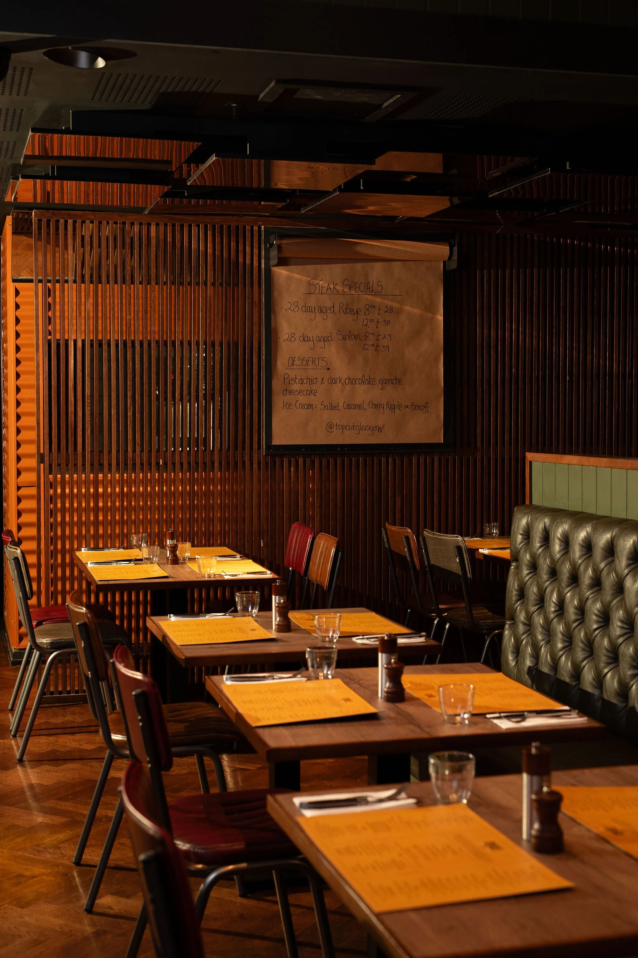 Empty restaurant with wooden tables, chairs, and menu placemats. A large brown paper menu board hangs on the wall.