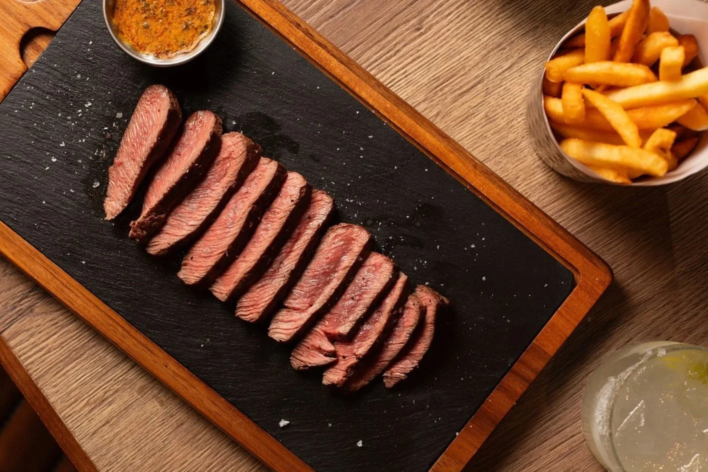 Two plates of sliced cooked steak on black wooden boards, surrounded by side dishes including french fries, roasted vegetables, macaroni and cheese, and salads, with glasses of red wine on a wooden table.