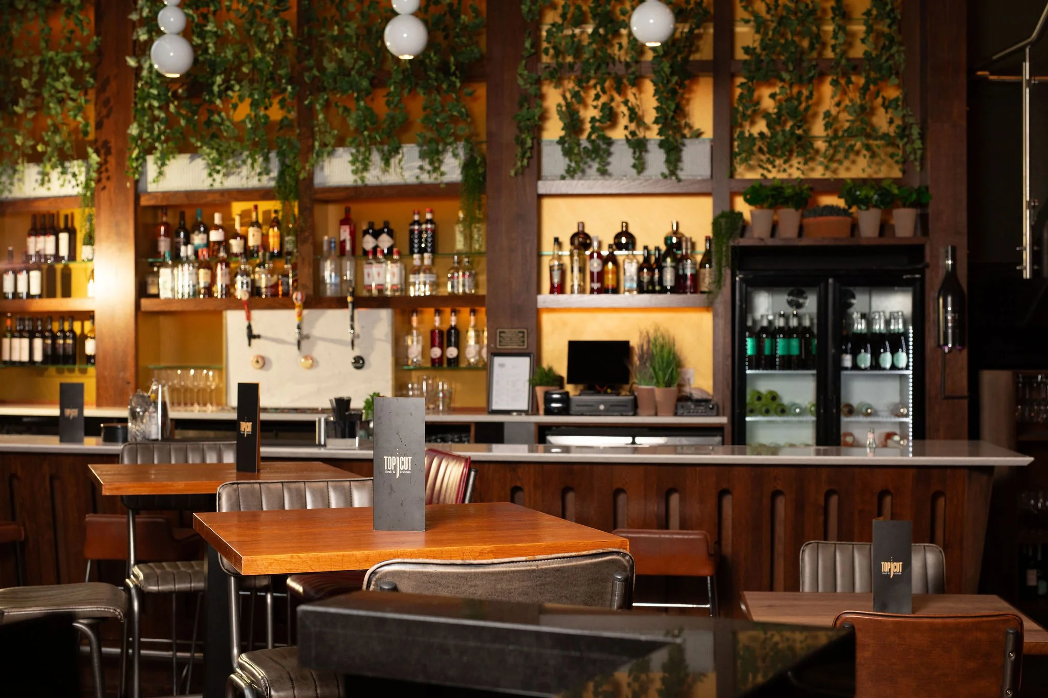 Empty bar and dining area with wooden tables, leather chairs, and a backlit bar shelf with various liquor bottles, hanging light fixtures, and green potted plants.