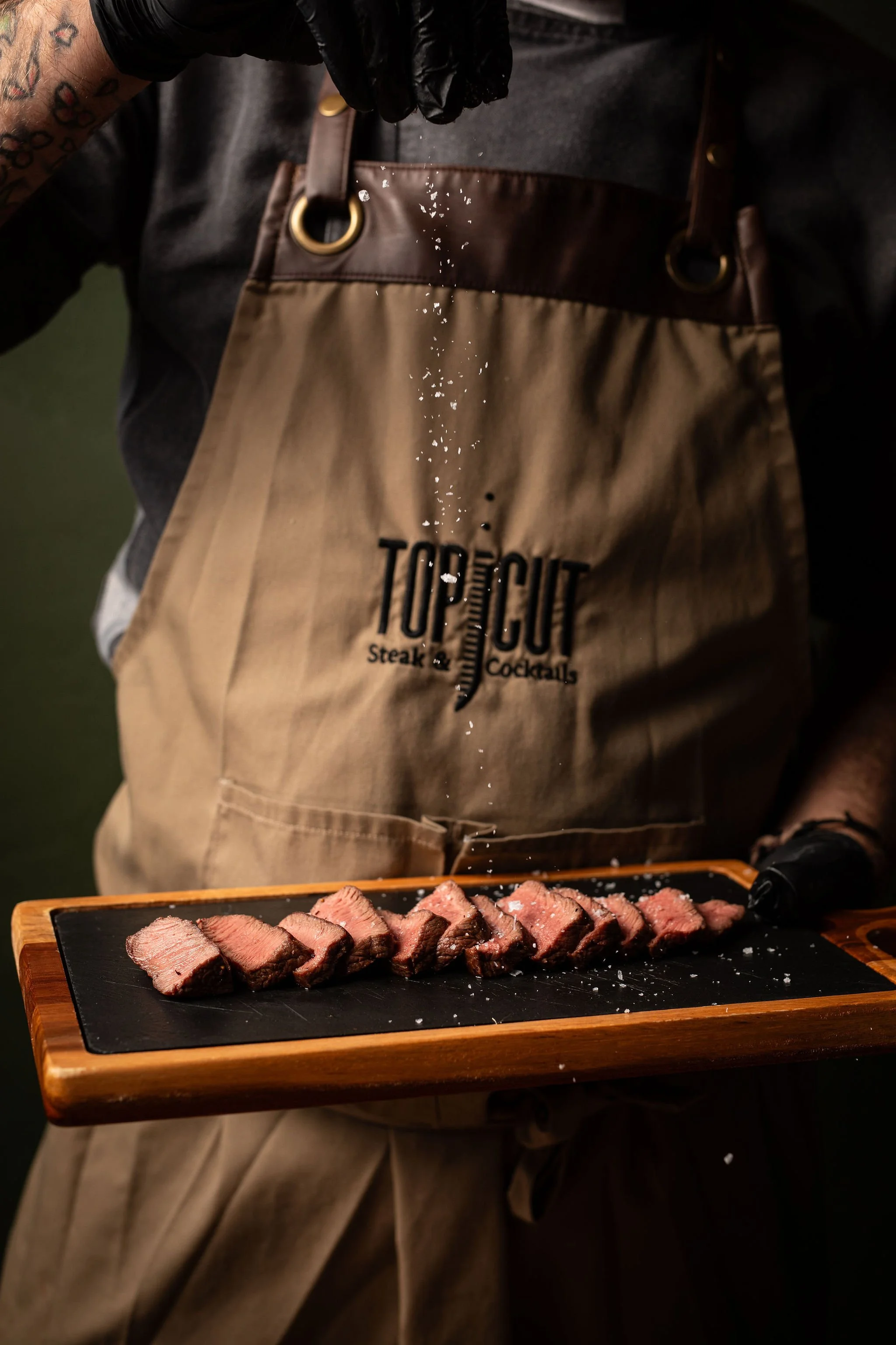 Person sprinkling salt on slices of cooked steak on a black cutting board, wearing a beige apron with 'Top Notch' text, over a dark shirt.