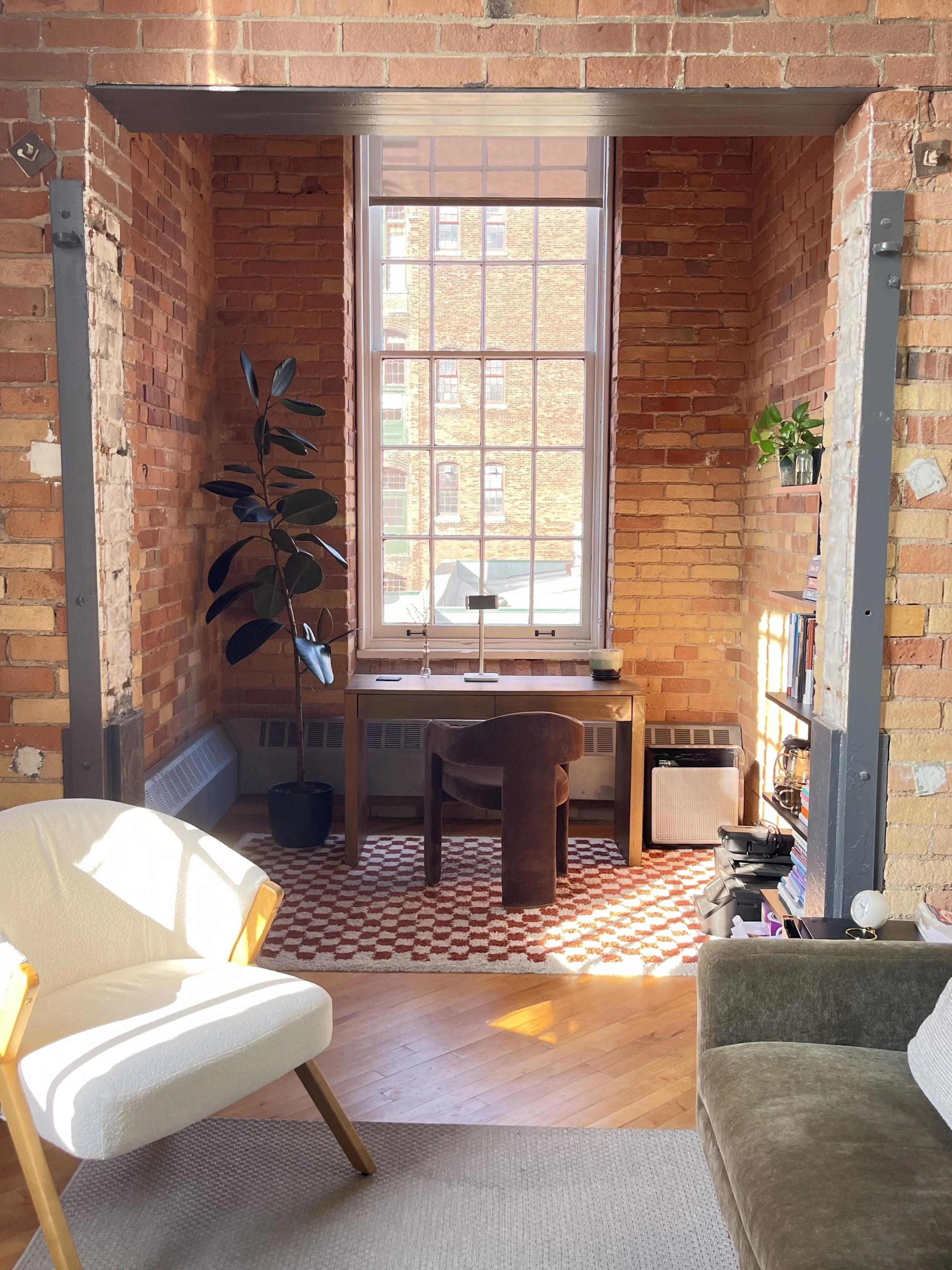 A cozy corner of a room with brick walls, a large window, and a view of neighboring brick buildings. The space features a small wooden desk with a chair, a potted plant, a bookshelf, and a patterned rug. There is sunlight casting shadows on the furniture.