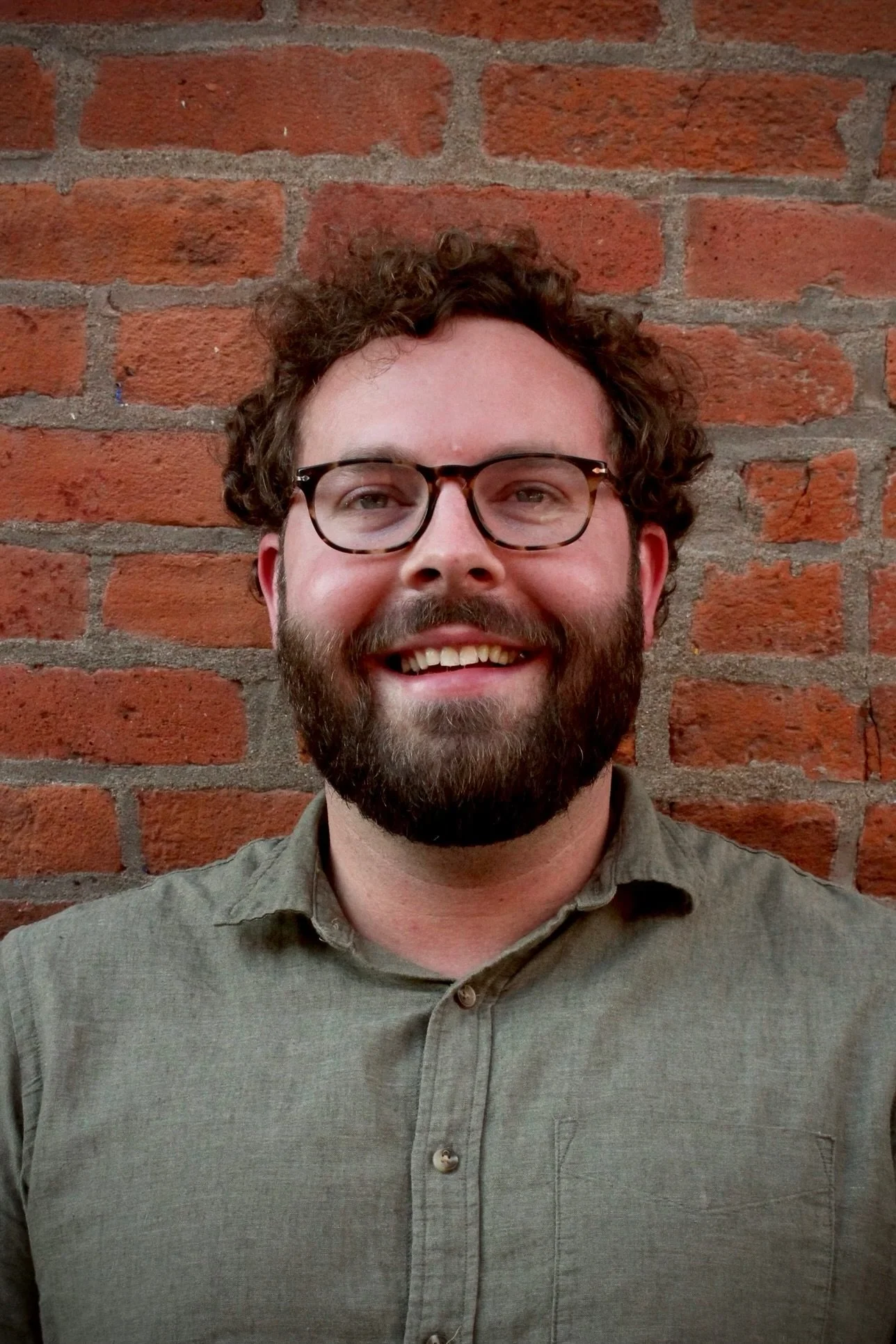 A man with curly hair, glasses, and a beard smiling in front of a brick wall.