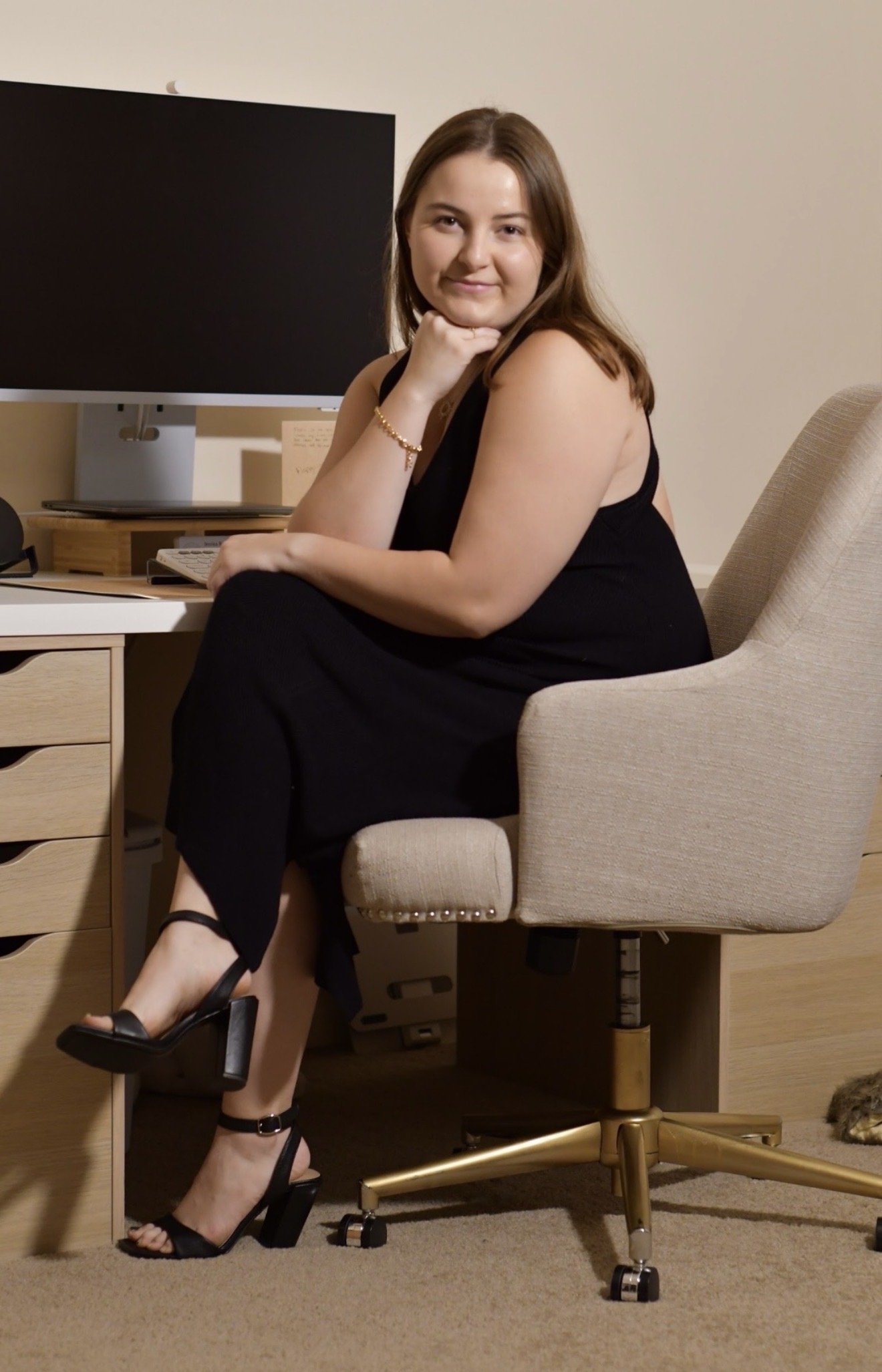 A woman with brown hair and light skin sitting in an office chair at a desk, wearing a black dress and black high-heeled sandals, with her right arm resting on her knee and her chin on her hand, smiling at the camera.
